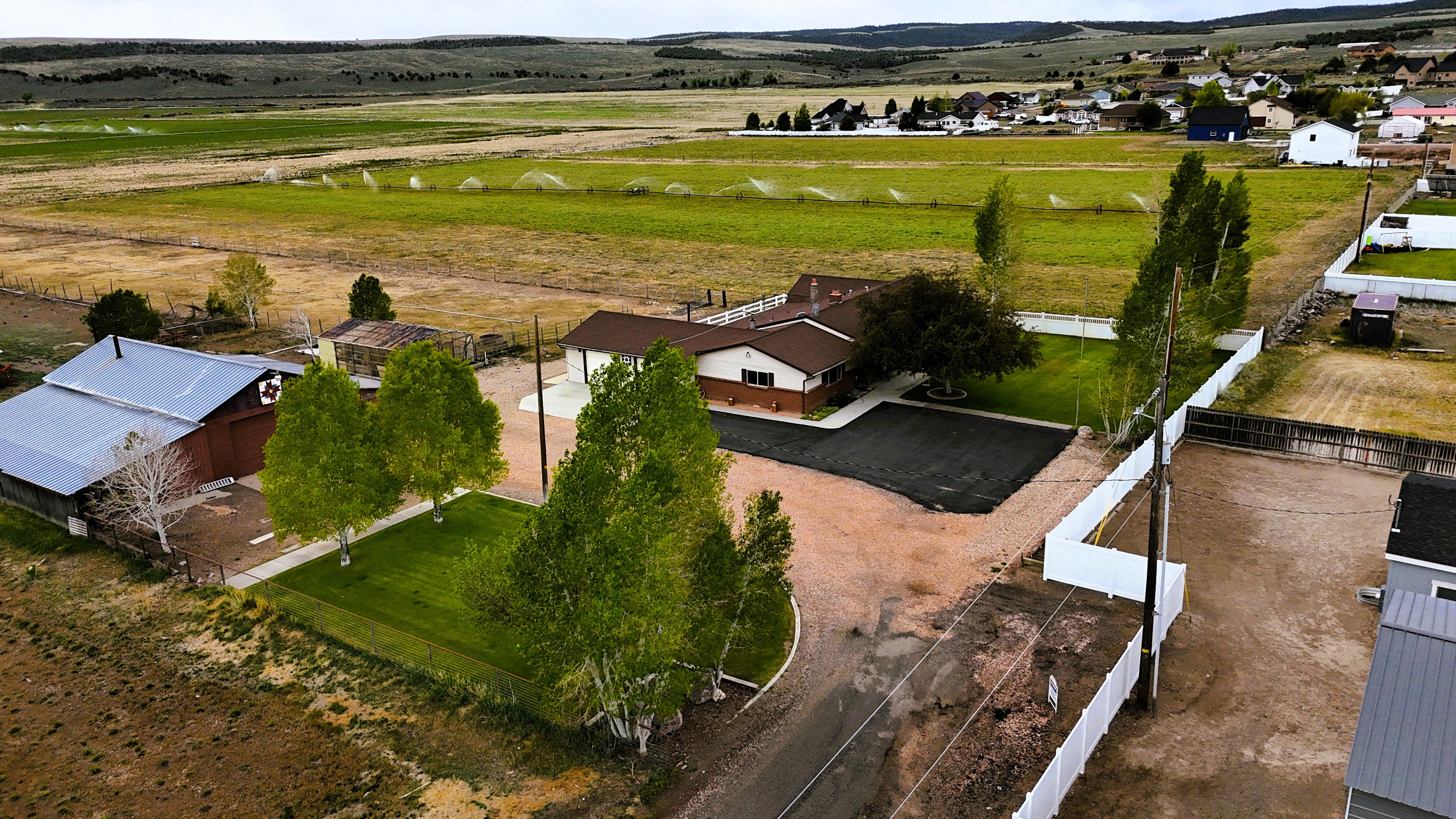 Expansive property featuring a ranch-style residence with a brown shingle roof, a large unpaved drive, manicured green lawns, a white perimeter fence, and mature trees