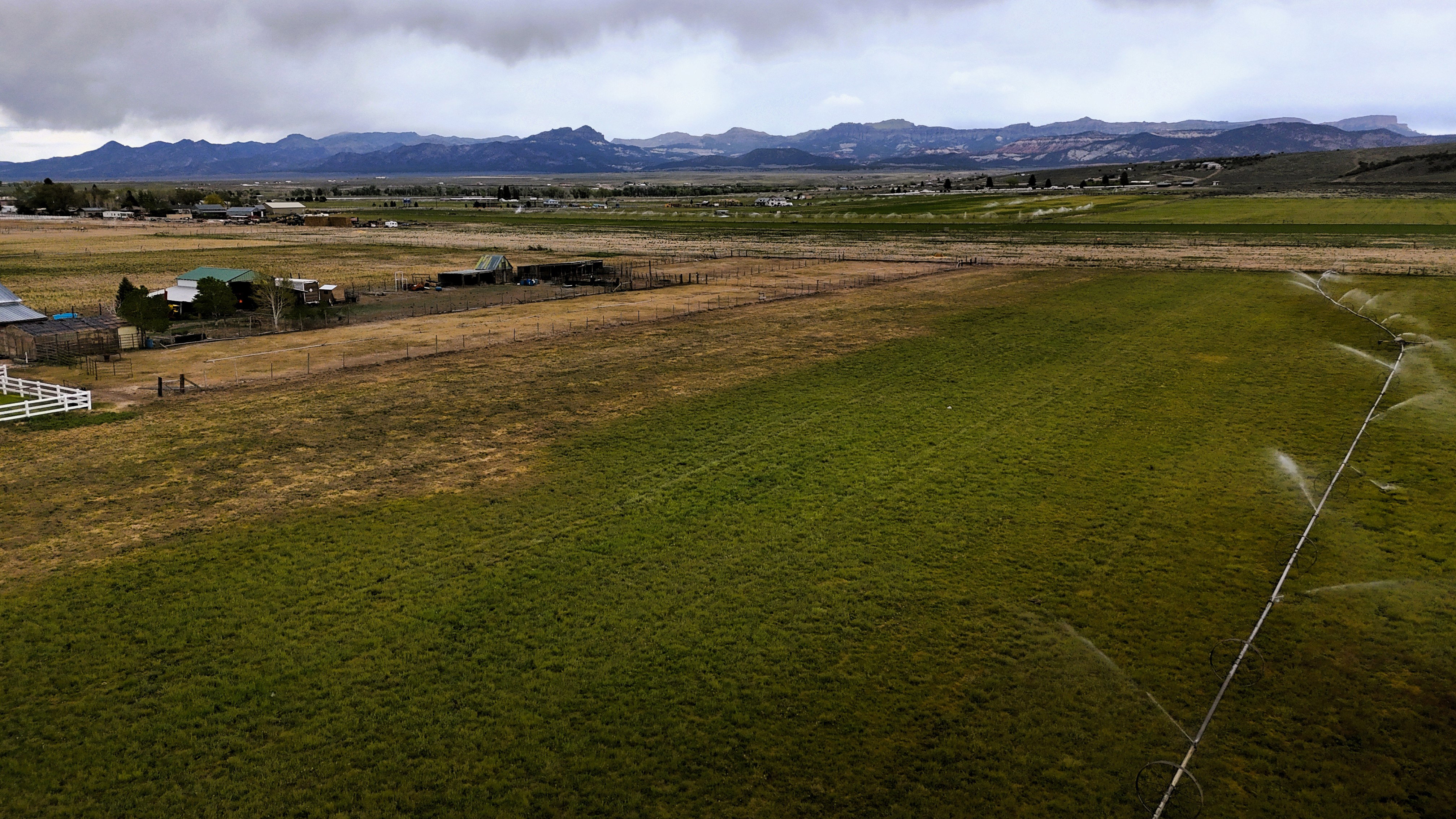 Expansive agricultural land featuring a green cultivated field with active irrigation, brown pasture areas, and a distant mountain range