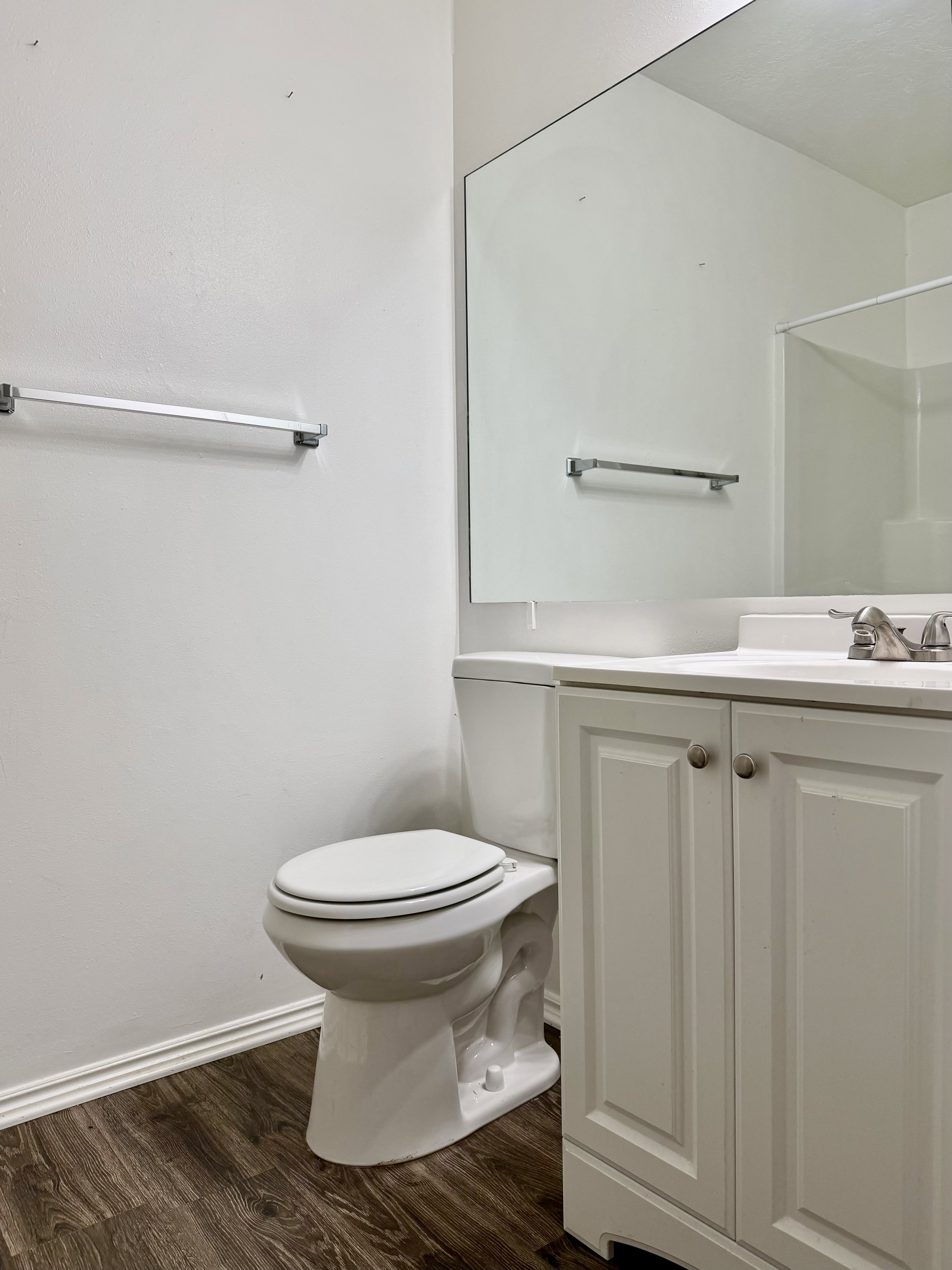 Bathroom featuring wood-finish flooring, a white vanity with paneled doors, a white countertop, a white toilet, and a large wall-mounted mirror
