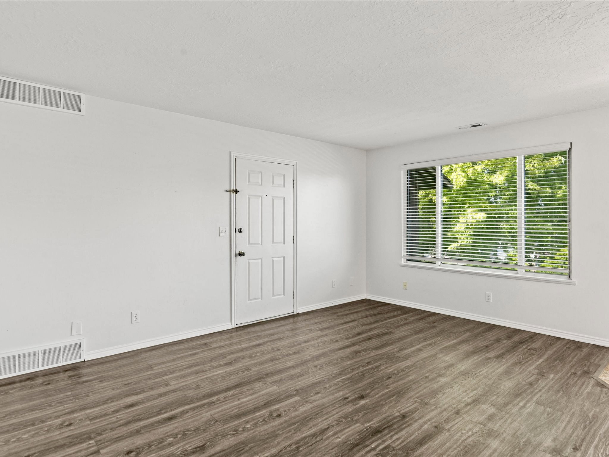 Spacious room with wood-finish flooring, white baseboards, and a large window featuring horizontal blinds