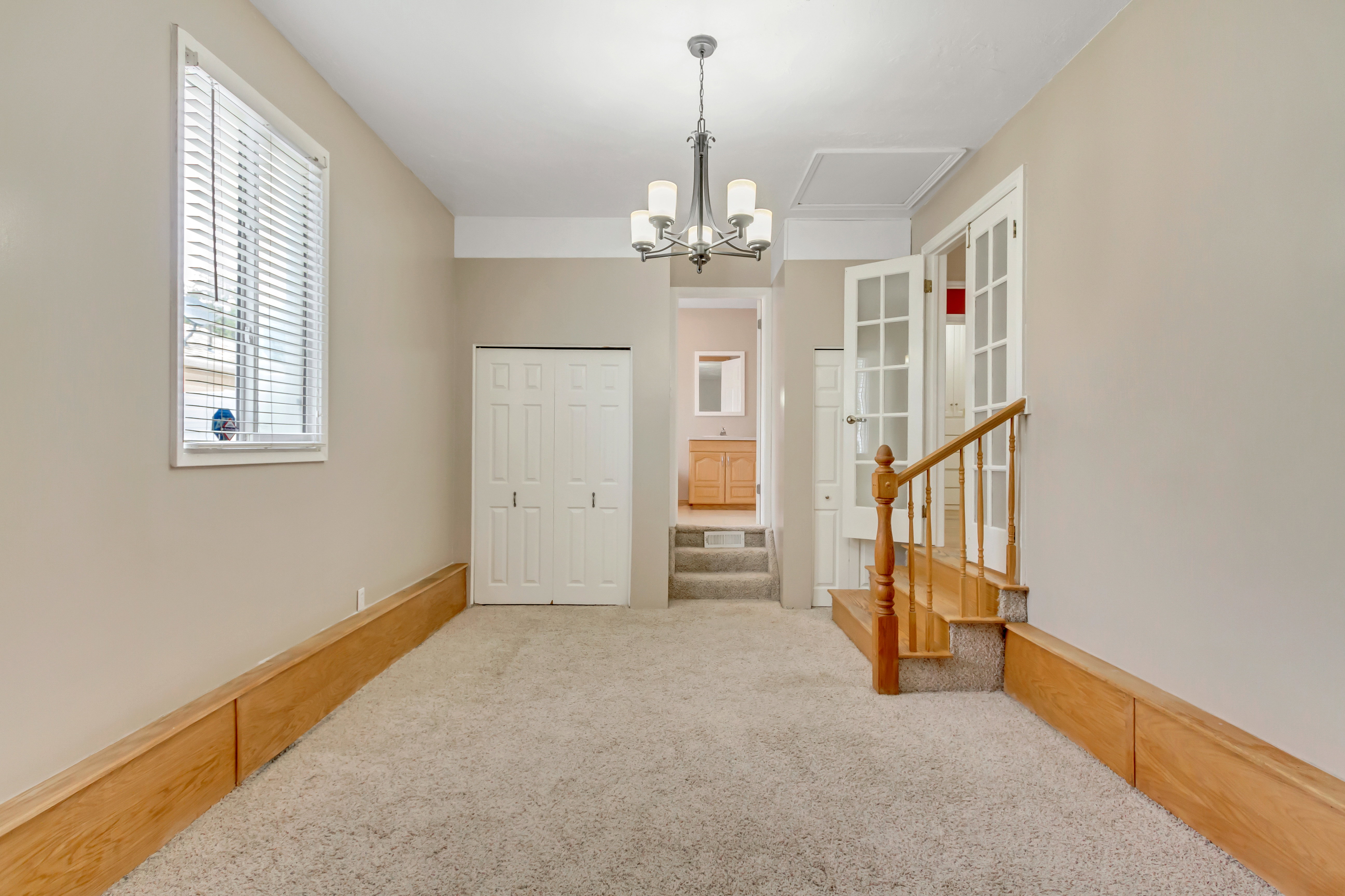 Carpeted room featuring a window with blinds, bi-fold doors, a chandelier, and a wood-finish staircase with a railing leading to French doors
