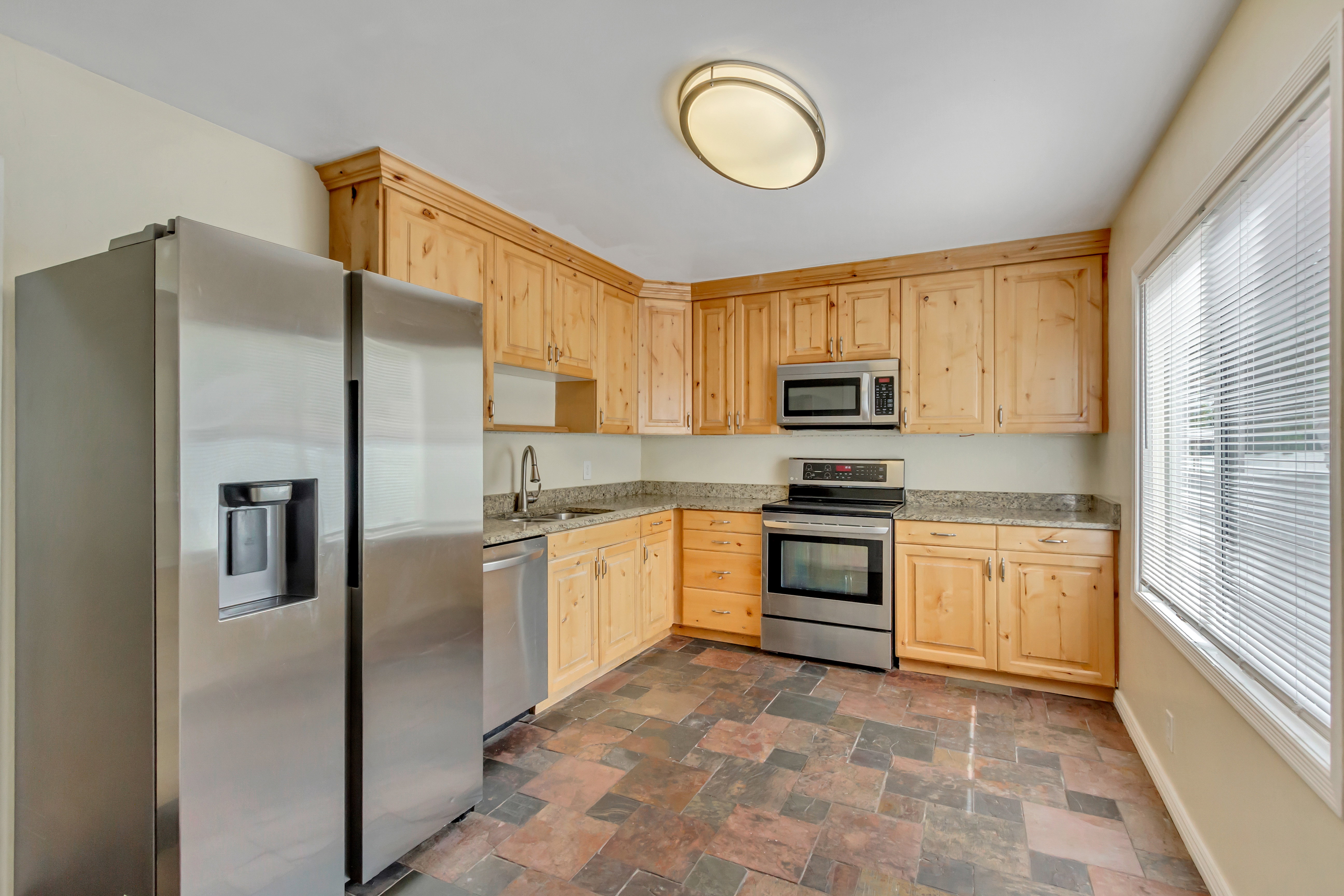 Kitchen featuring wood cabinetry, stainless steel appliances, speckled stone-finish countertops, and large format floor tiles
