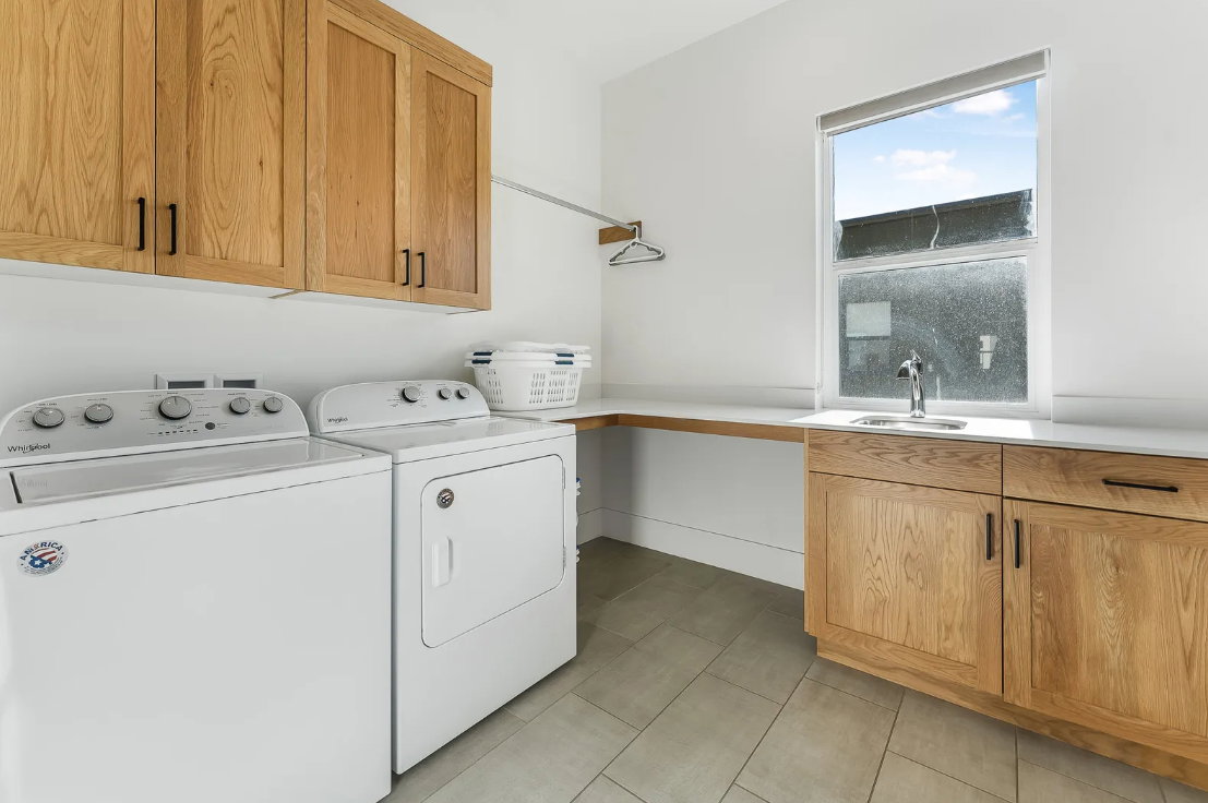 Laundry room featuring natural wood-finish cabinetry with matte black hardware, a built-in sink with a chrome faucet, and a large window