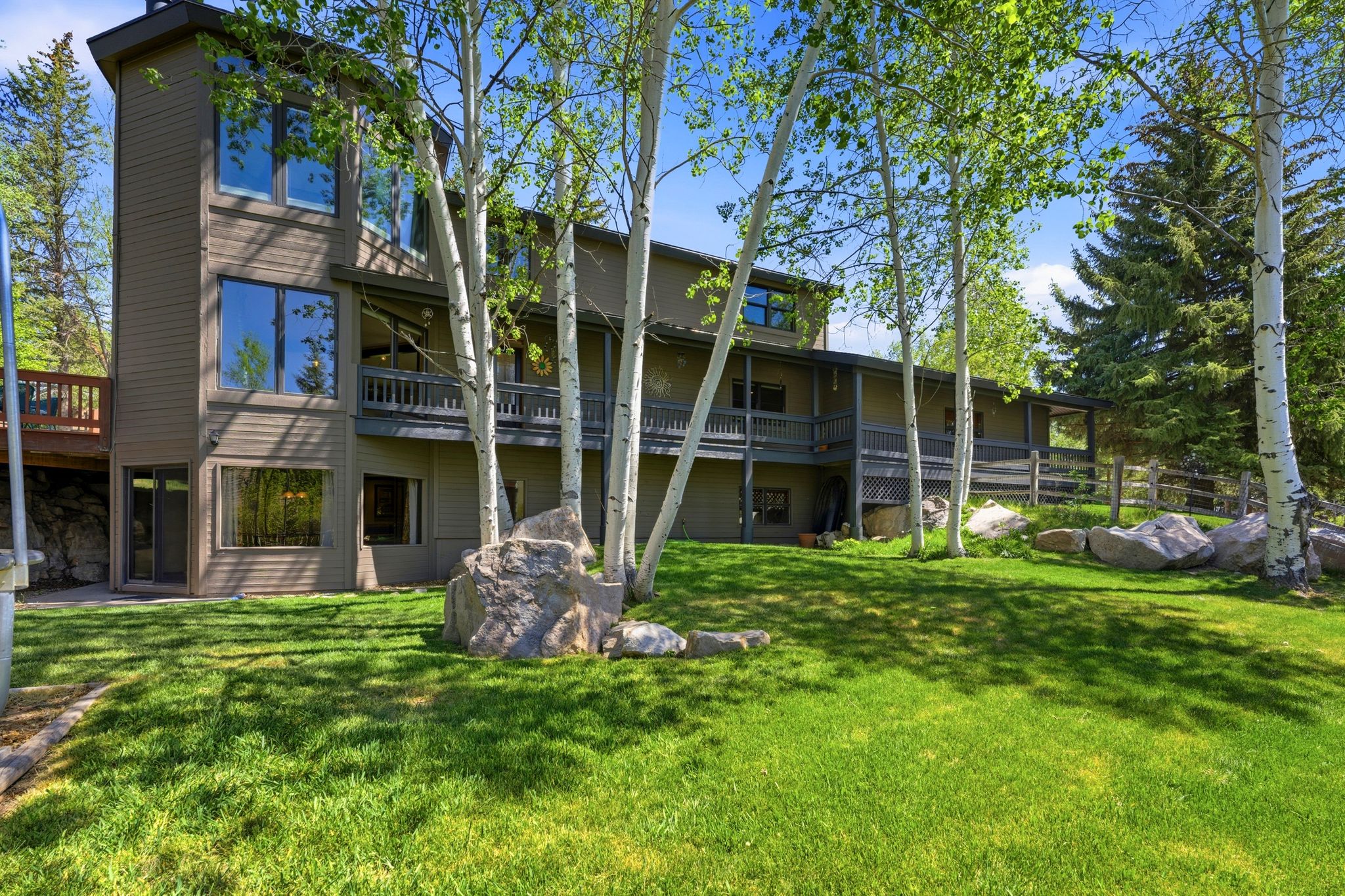 Multi-level home featuring extensive decking, a manicured lawn, mature trees, and natural rock landscaping