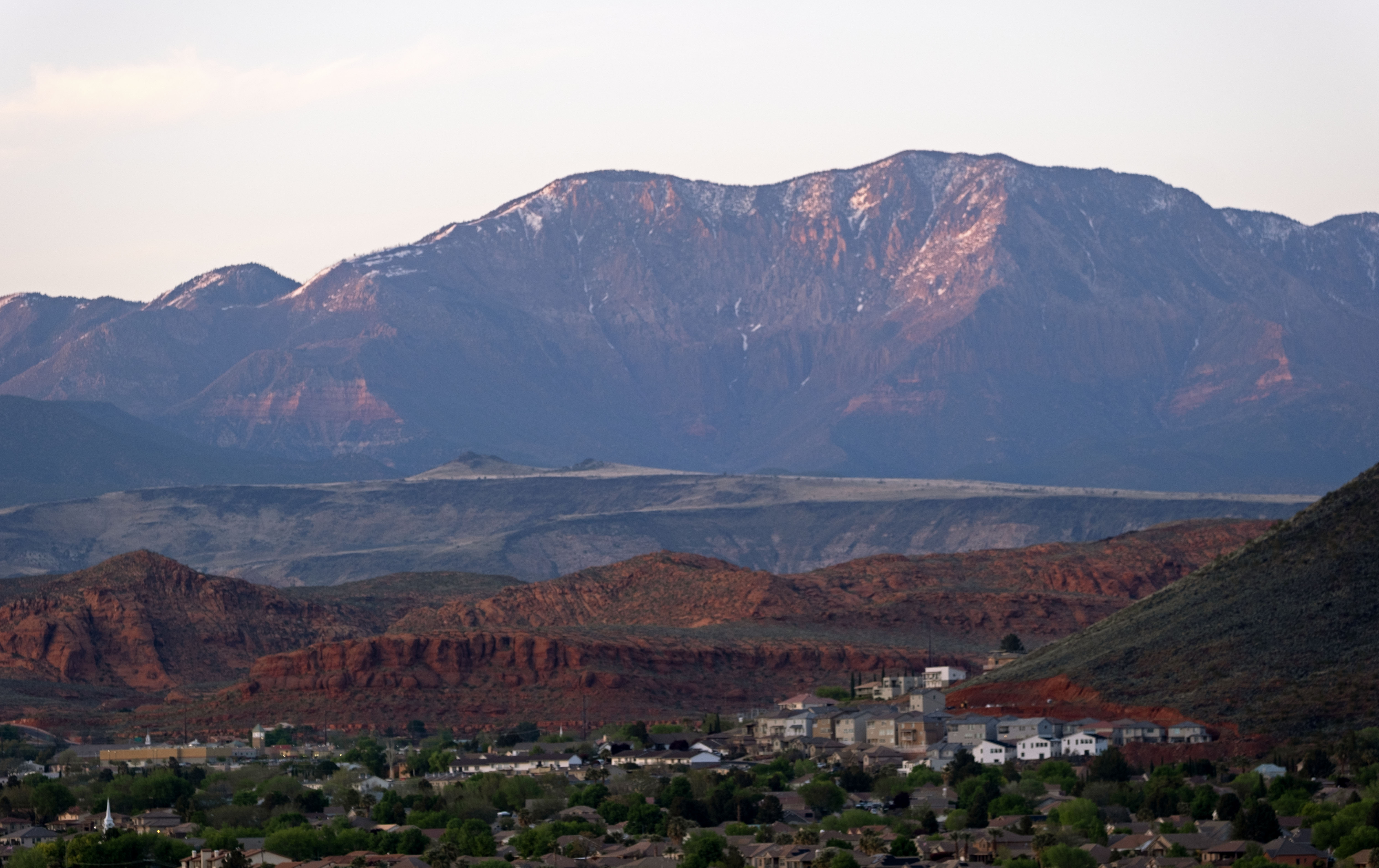 Mountainous landscape featuring snow-capped peaks, red rock formations, and a distant community