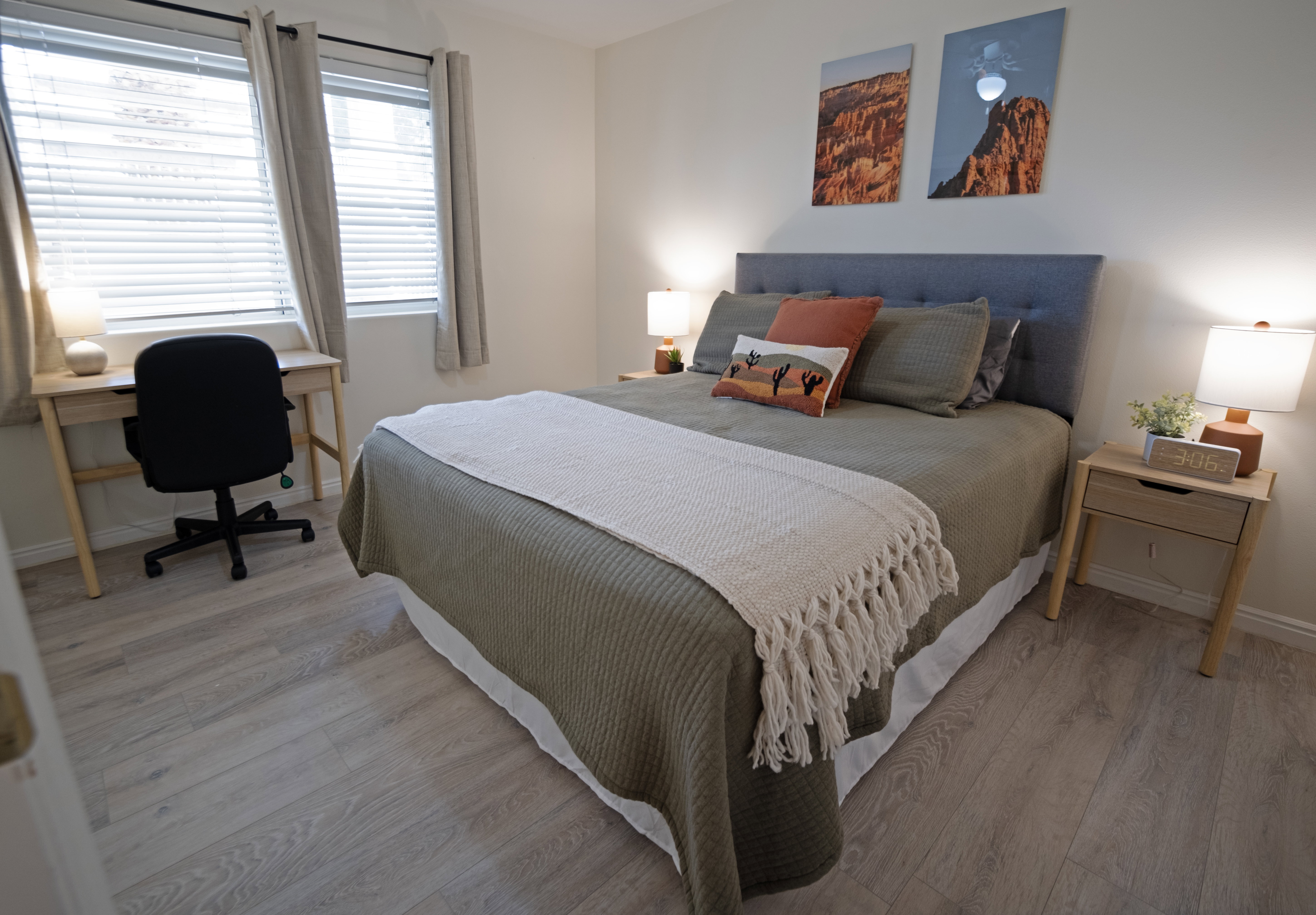 Bedroom featuring light wood-finish flooring, a tufted fabric headboard, two windows with blinds and curtains, and a built-in desk area
