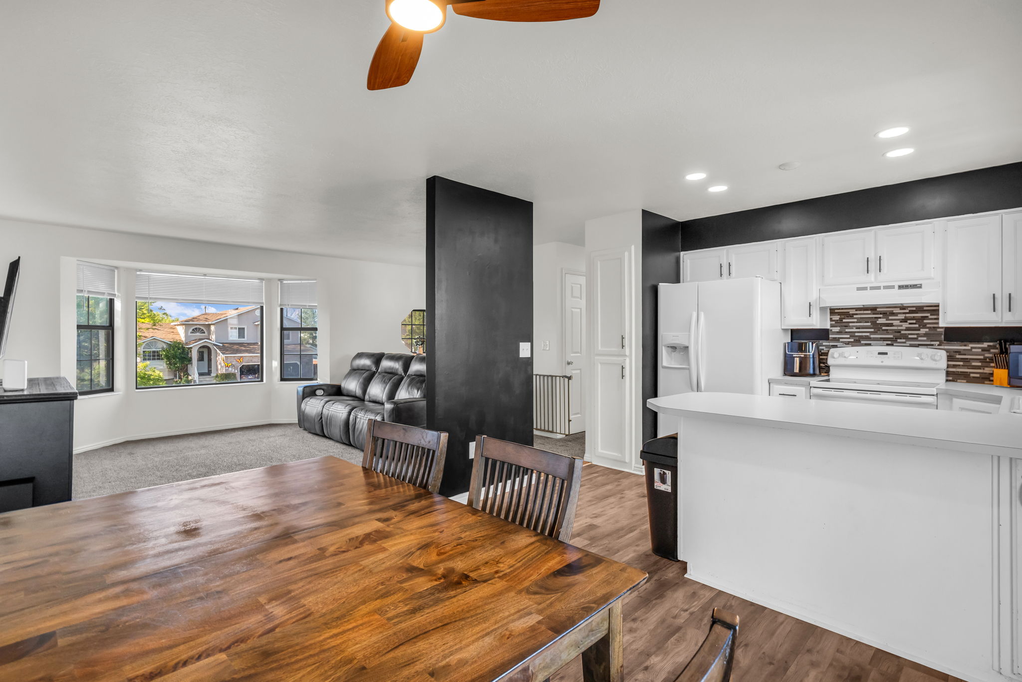 Open-concept living space featuring a central dining area with wood-finish flooring, a kitchen with white cabinetry and a tile backsplash, and a carpeted living area with a large bay window