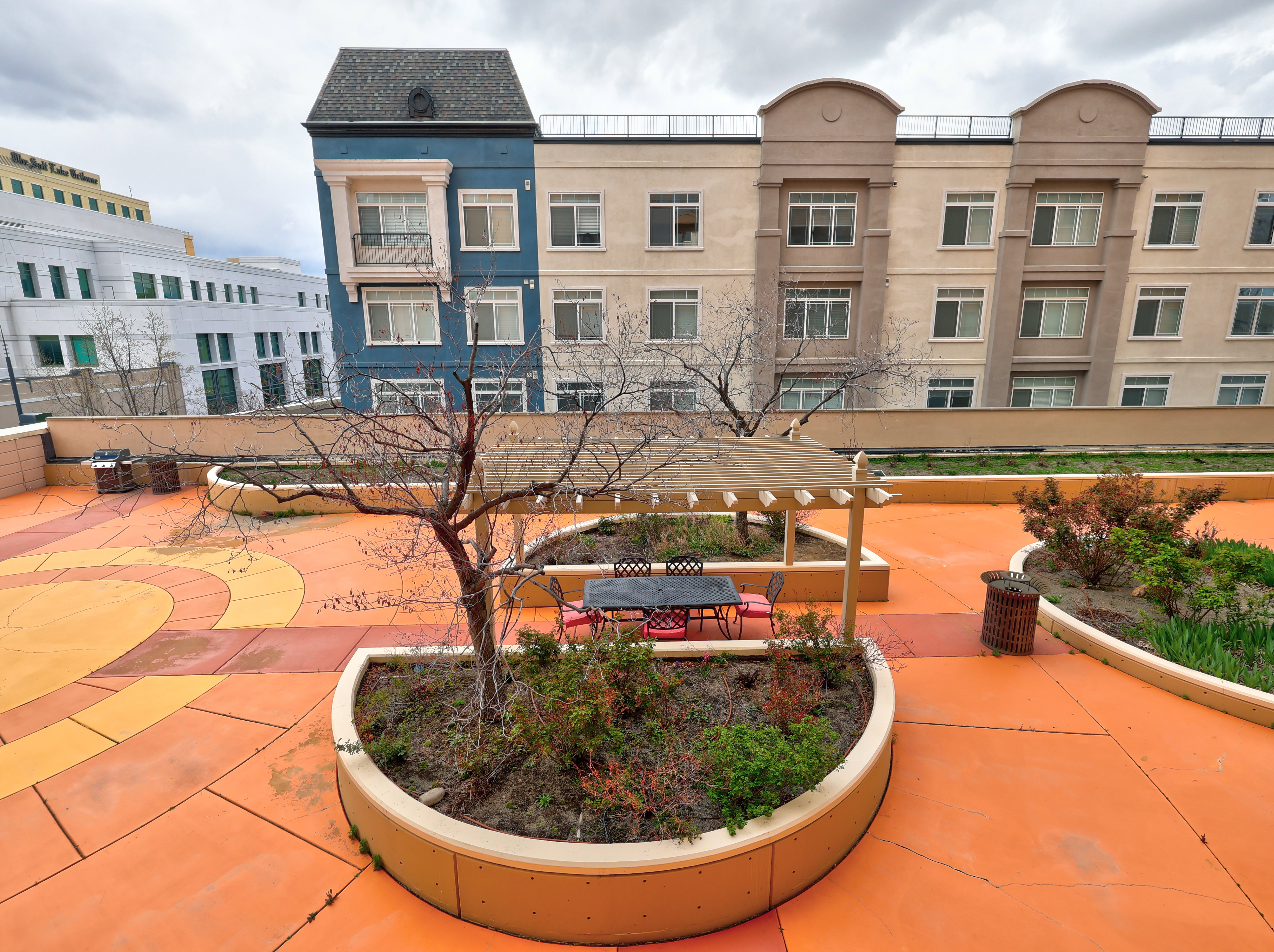Rooftop patio featuring expansive orange and yellow patterned flooring, multiple built-in planter beds with mature trees and shrubs, and a shaded pergola