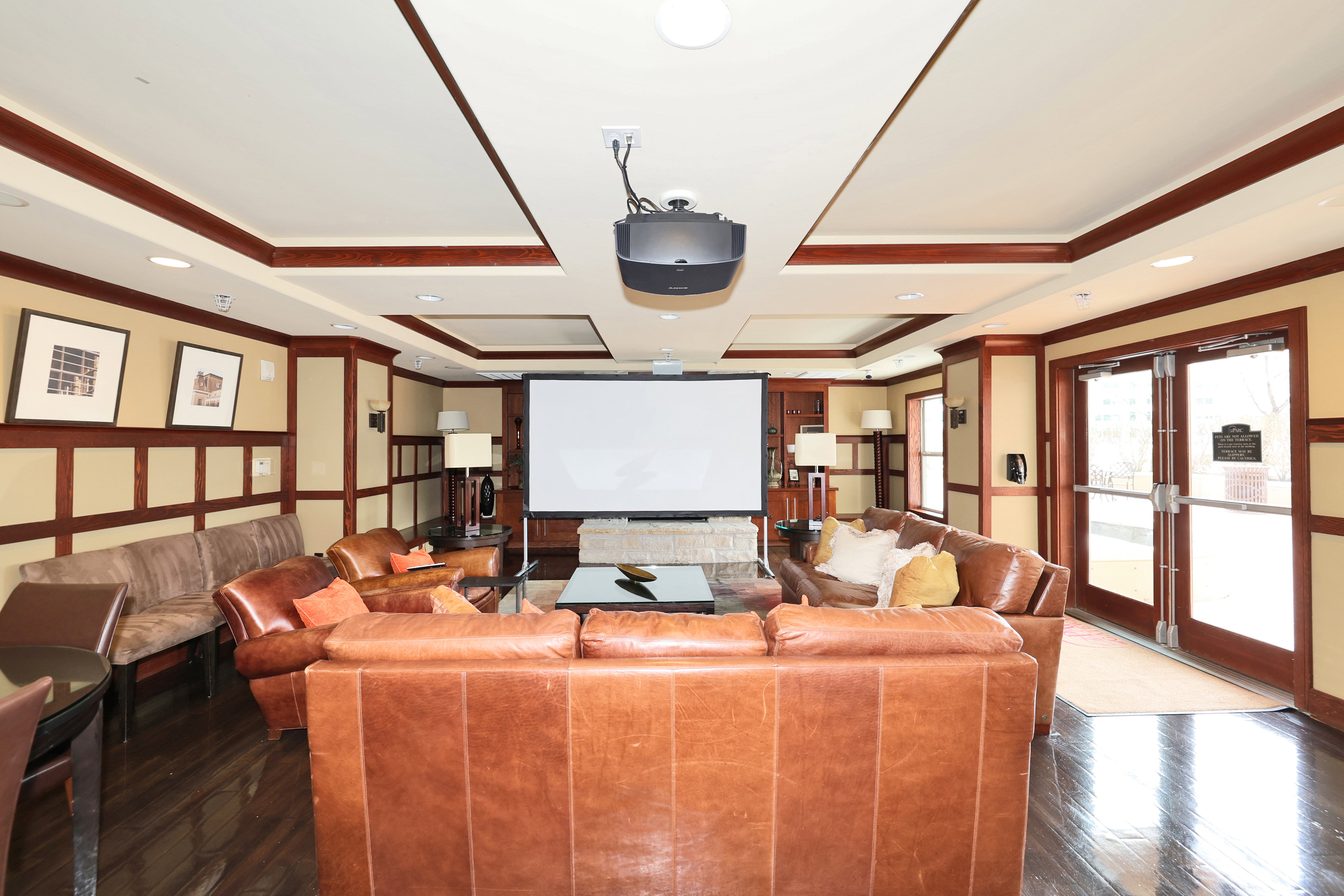 Spacious entertainment area featuring a ceiling-mounted projector, large screen, tray ceilings with recessed lighting, dark wood-finish flooring, and wainscoting