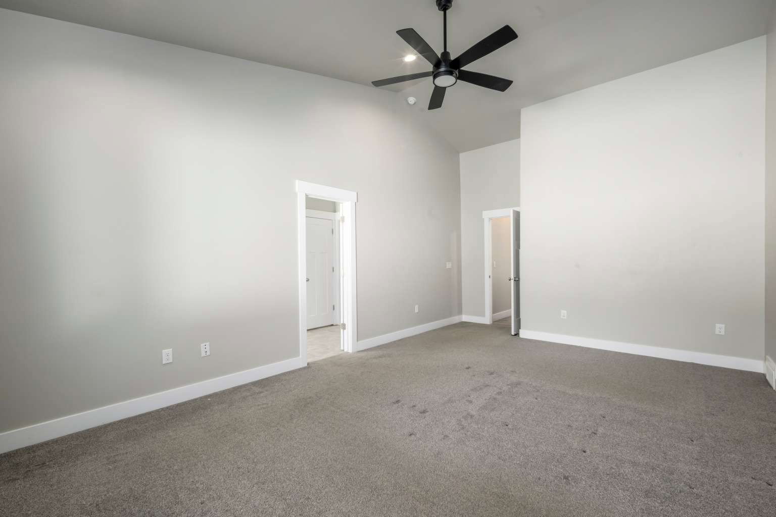 Spacious interior room featuring vaulted ceilings, neutral gray wall paint, dark carpet flooring, and a modern black ceiling fan