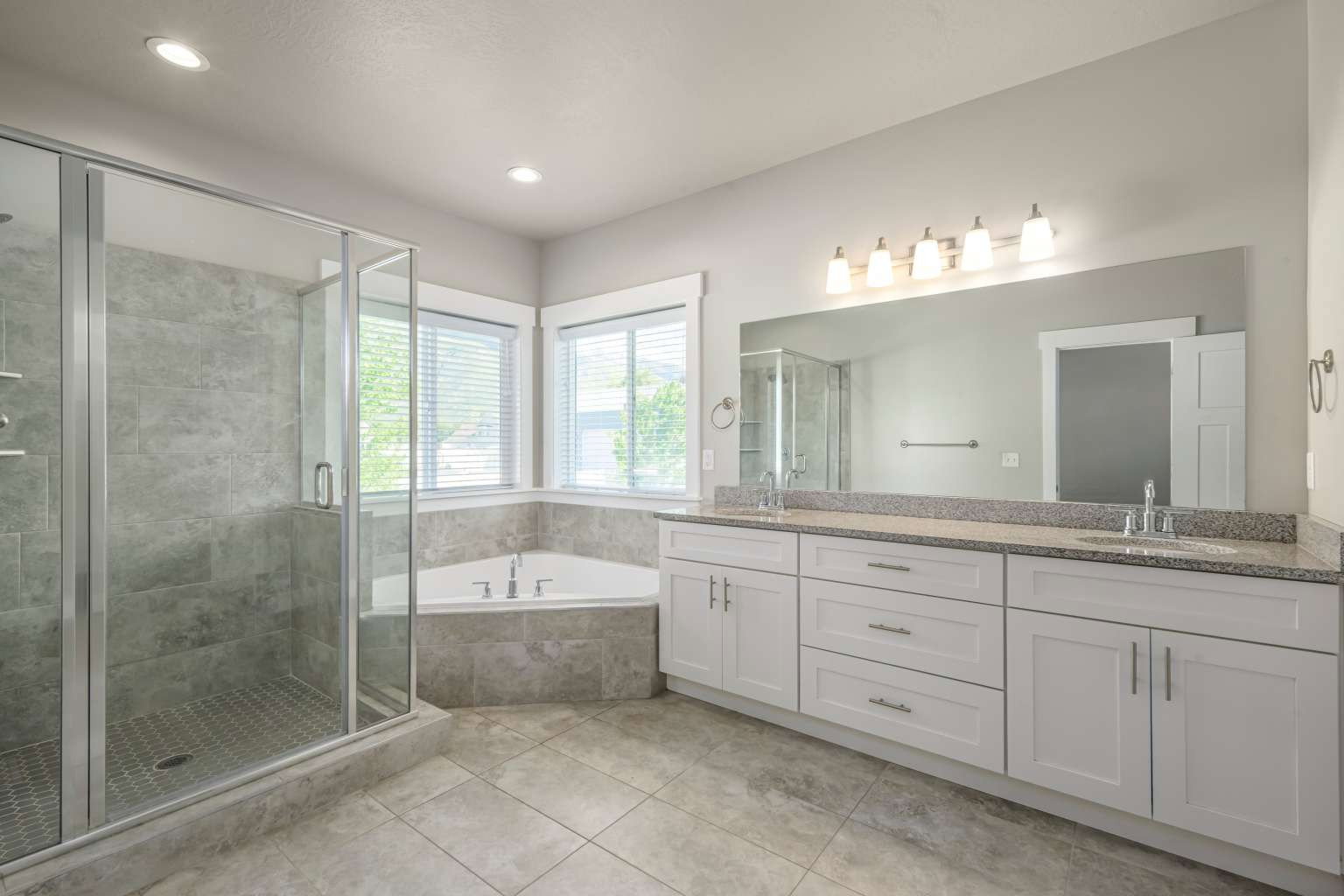 Spacious bathroom featuring a glass-enclosed shower with tiled walls, a corner soaking tub, dual vanity with granite countertops, and light-colored cabinetry