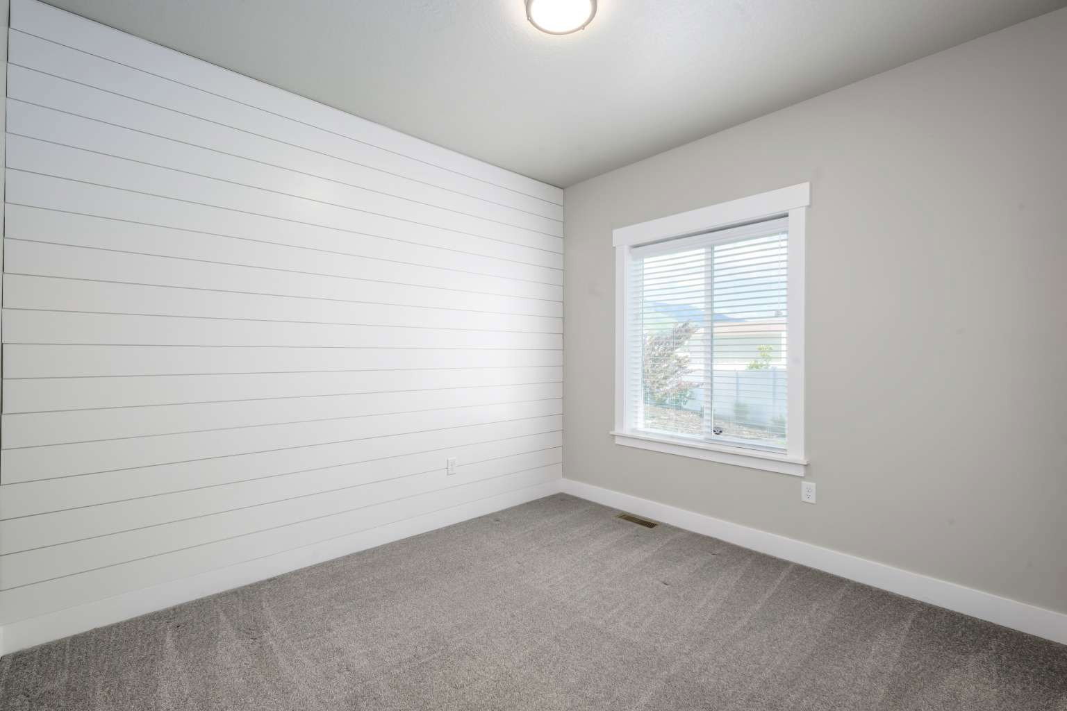 Room featuring a white shiplap accent wall, neutral painted walls, gray carpet flooring, and a single window with white trim and blinds