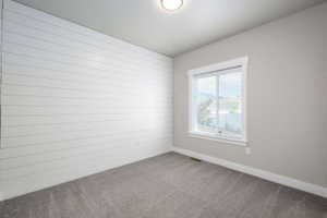 Room featuring a white shiplap accent wall, neutral painted walls, gray carpet flooring, and a single window with white trim and blinds