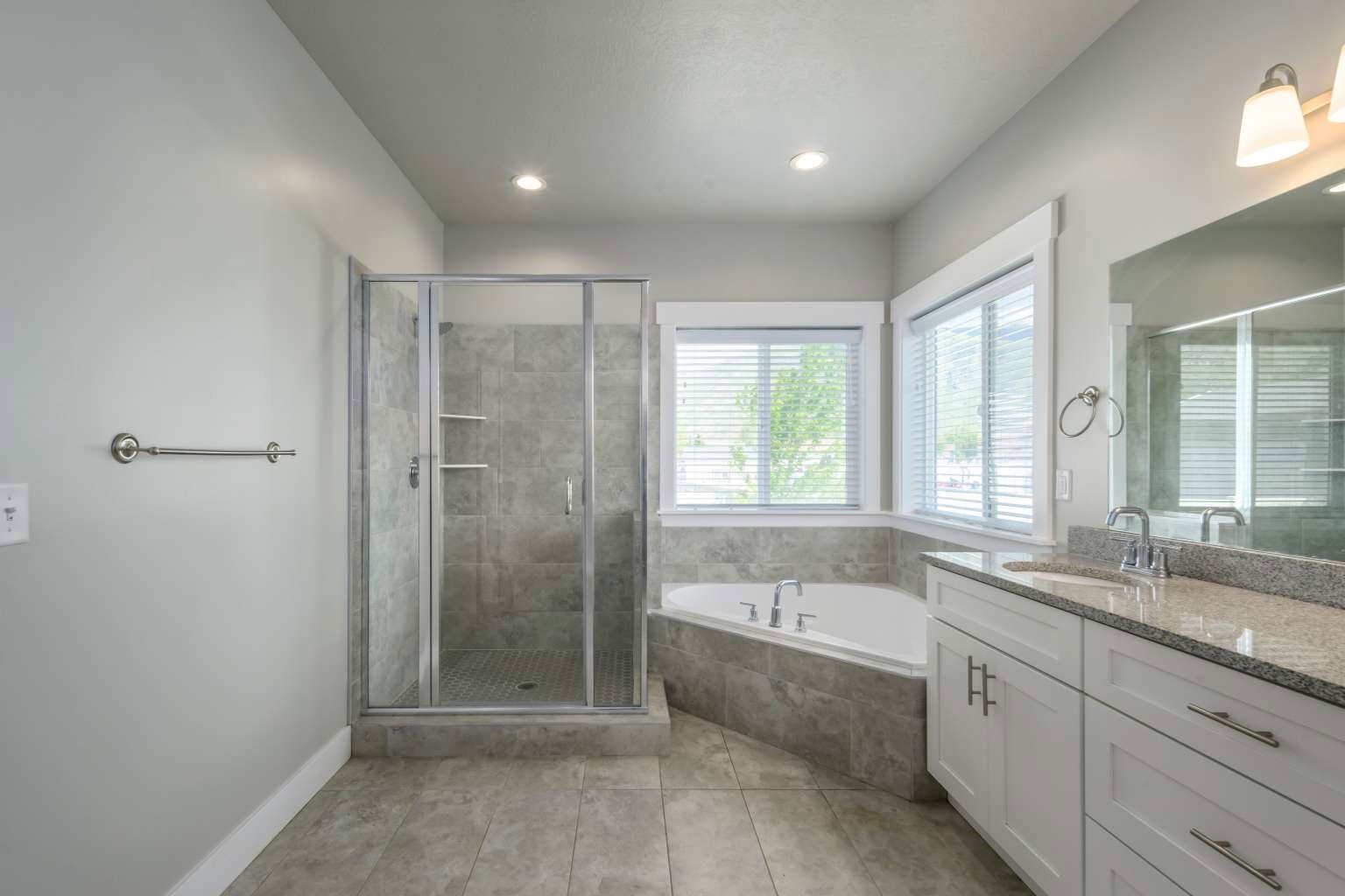 Spacious bathroom featuring a glass-enclosed shower, a corner built-in tub, a double vanity with stone countertop, and tile flooring