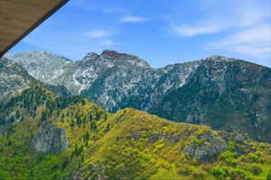 Expansive mountain landscape featuring varied elevations, vibrant green foliage, rocky outcrops, and snow-capped peaks against a clear blue sky