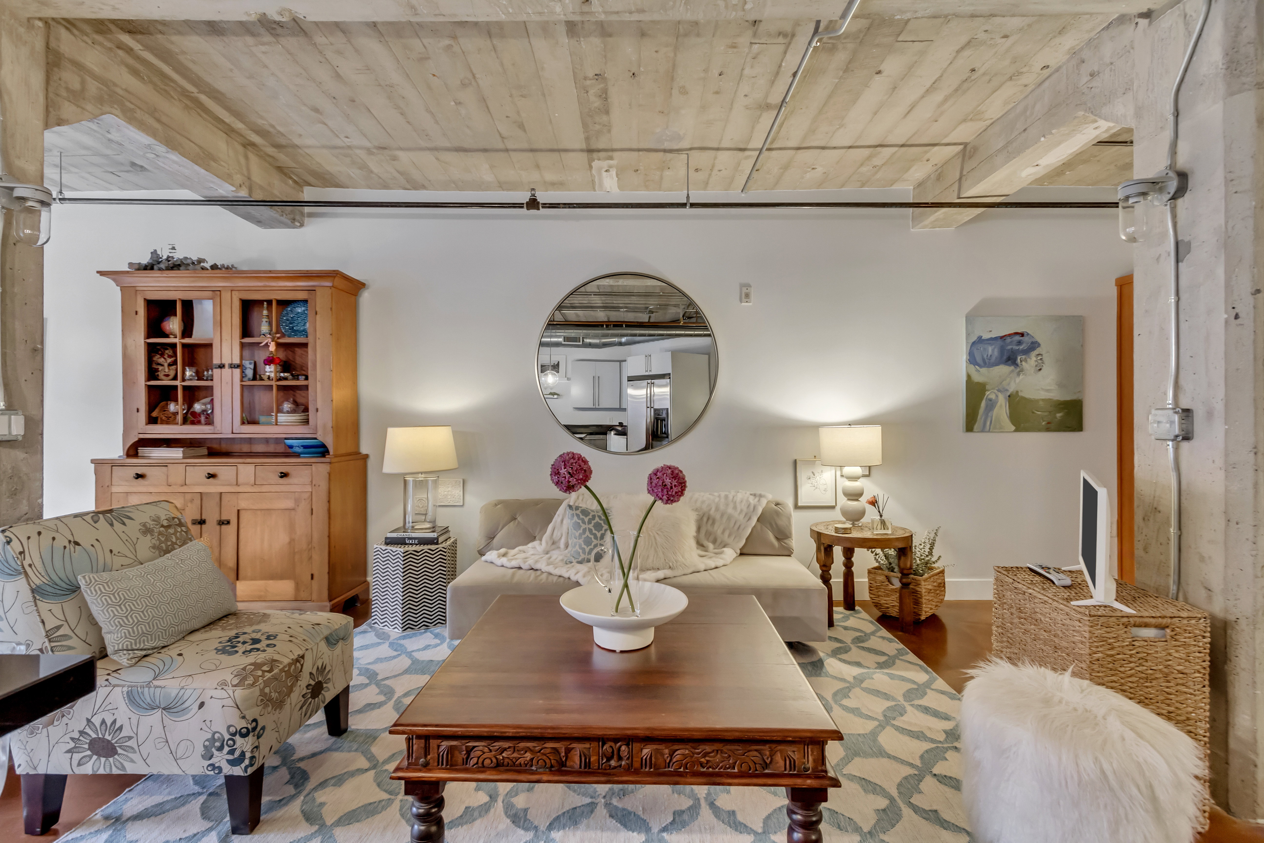 Spacious room featuring exposed concrete ceilings with visible wood beams, industrial-style conduit lighting, and light hardwood flooring partially covered by an area rug
