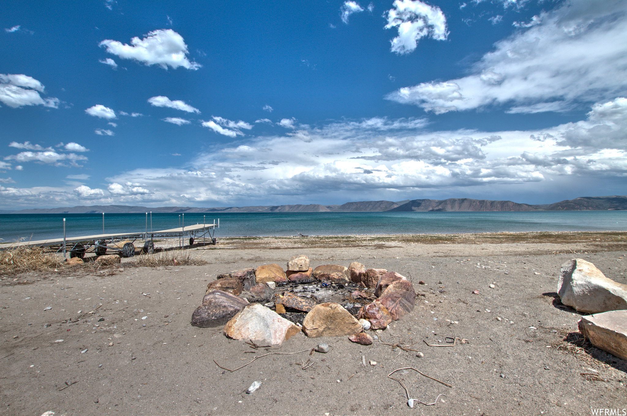 Lakeside property featuring a stone-lined fire pit, a private dock extending into the water, and expansive water frontage with mountain scenery