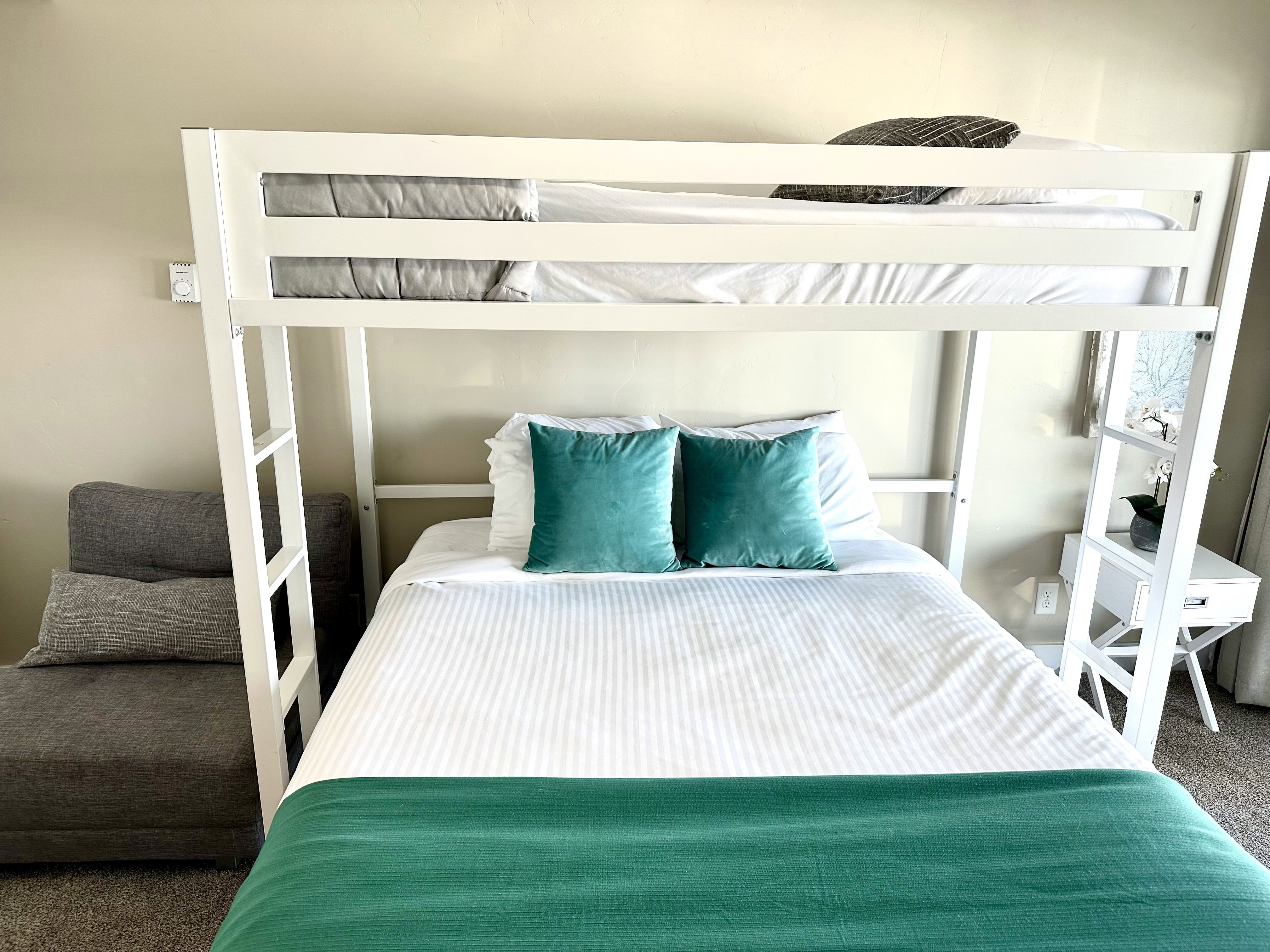 White bunk bed frame with integrated ladder, light beige wall paint, light-colored carpet, and a white side table with a drawer
