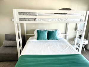 White bunk bed frame with integrated ladder, light beige wall paint, light-colored carpet, and a white side table with a drawer
