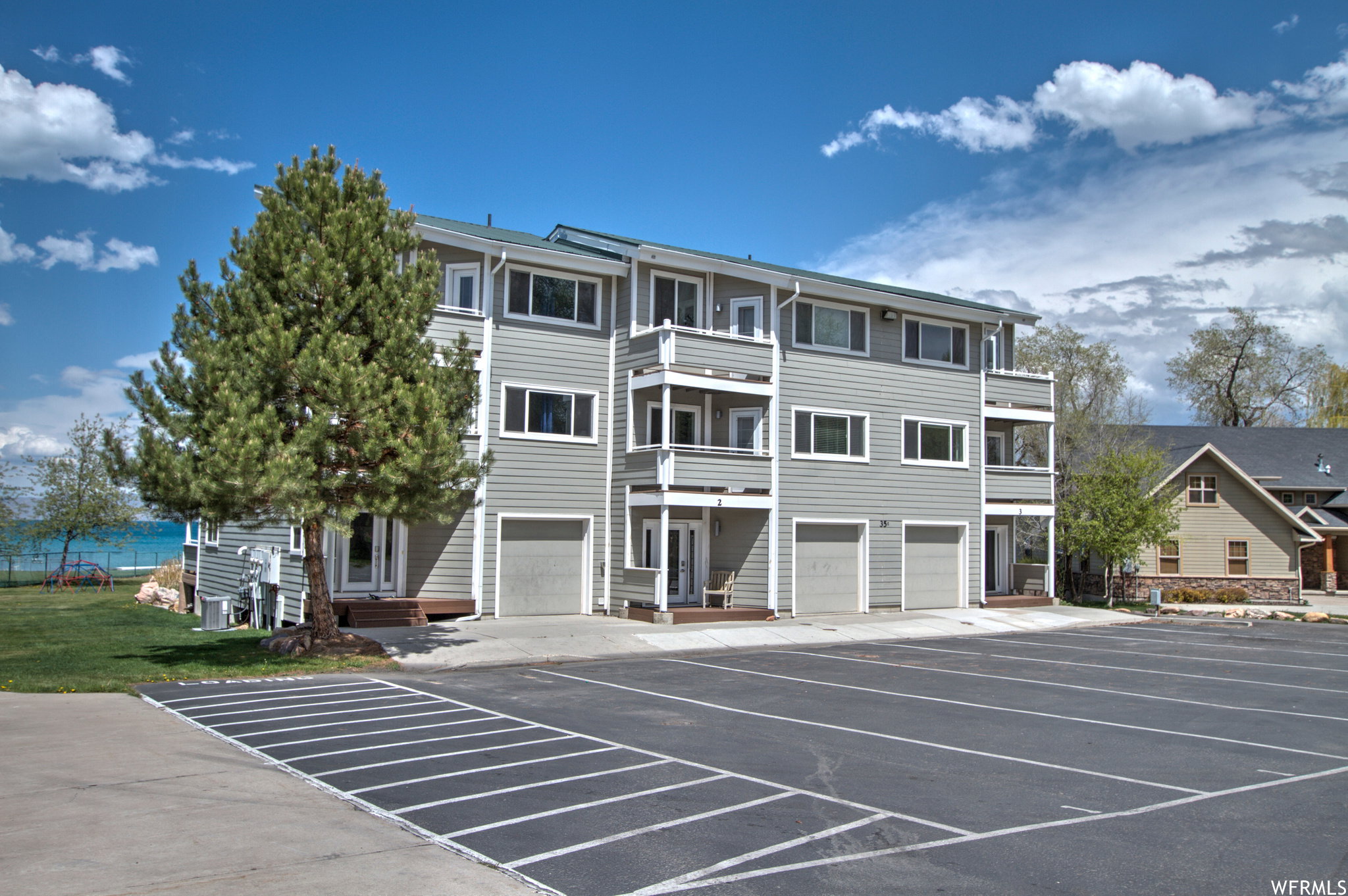Multi-story building featuring horizontal siding, multiple balconies, individual garage doors, and a paved parking area