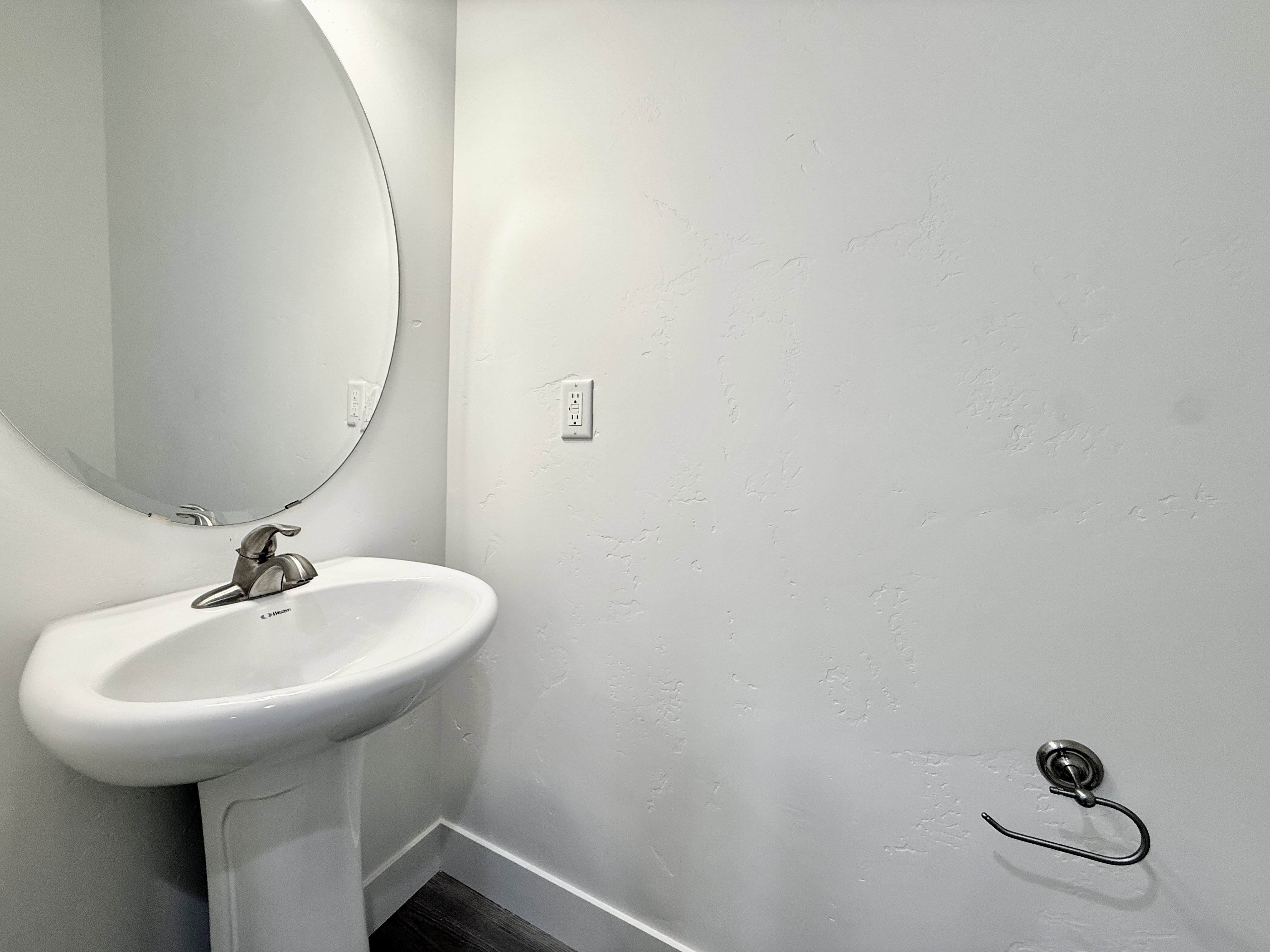 Pedestal sink with a polished chrome faucet, circular wall-mounted mirror, and dark wood-finish flooring