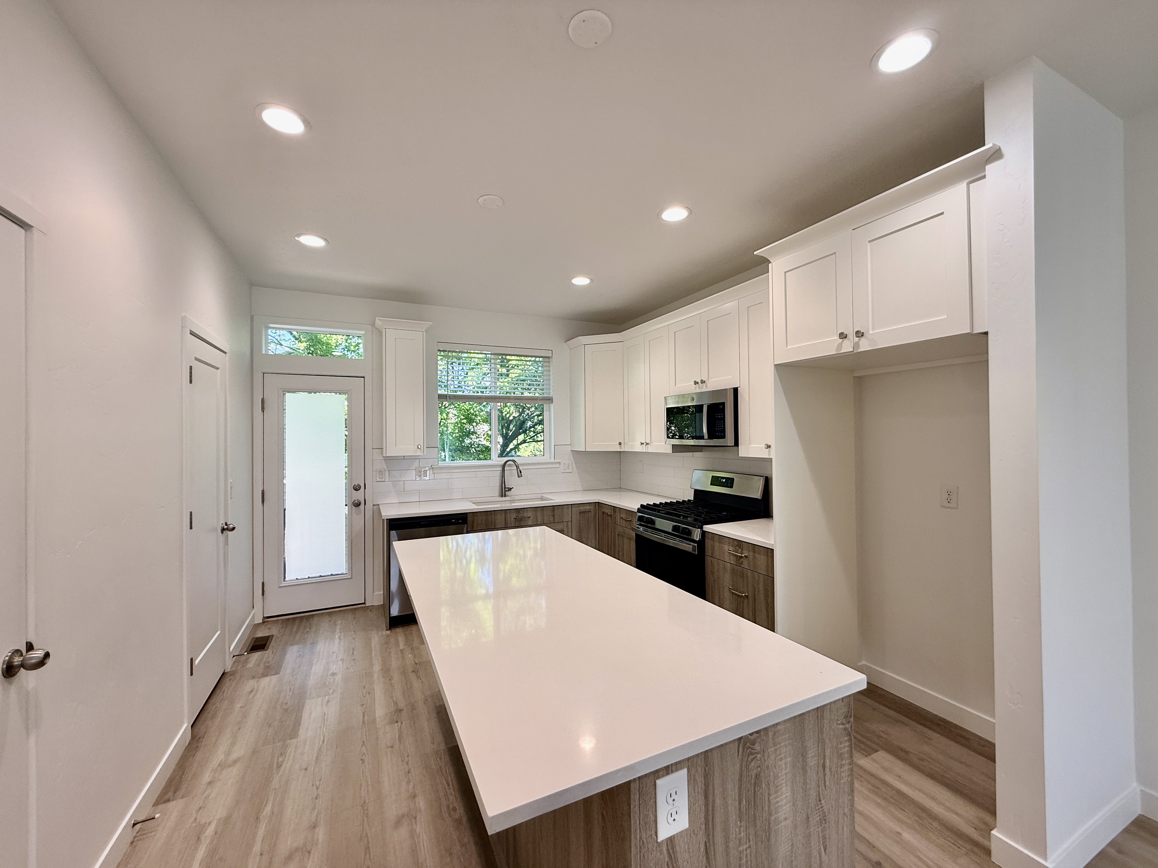 Modern kitchen featuring a large central island with a solid surface countertop, wood-finish flooring, white upper cabinetry, and stainless steel appliances including a gas range and microwave