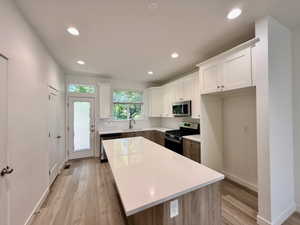 Modern kitchen featuring a large central island with a solid surface countertop, wood-finish flooring, white upper cabinetry, and stainless steel appliances including a gas range and microwave