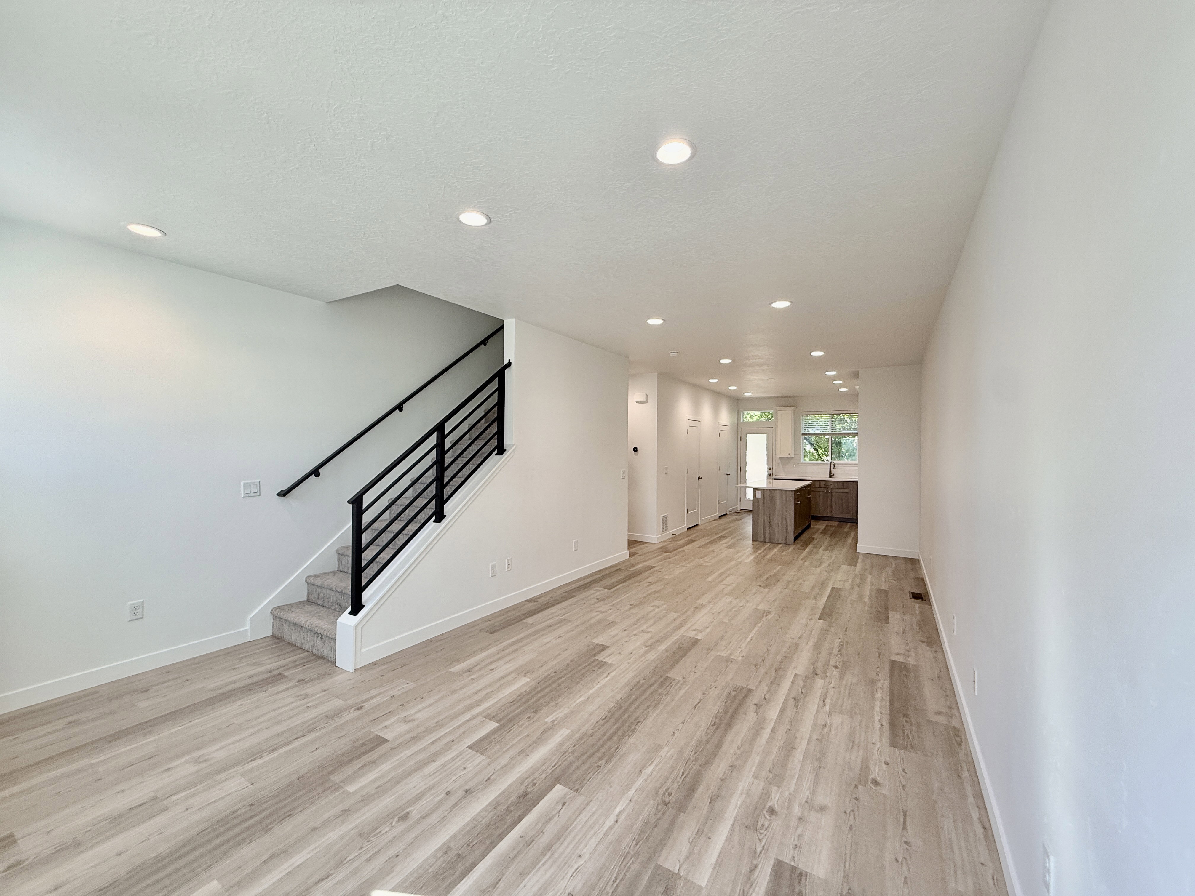 Expansive open-concept living area featuring wood-finish flooring, recessed lighting, a modern staircase with black metal railings, and crisp white walls