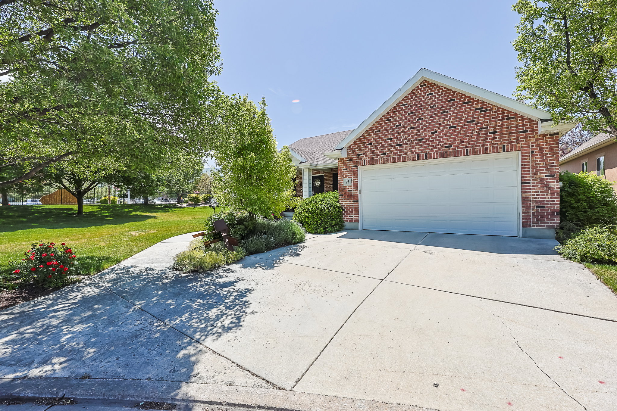 Brick facade residence featuring an attached garage with a white door, a concrete driveway, established landscaping, and mature trees