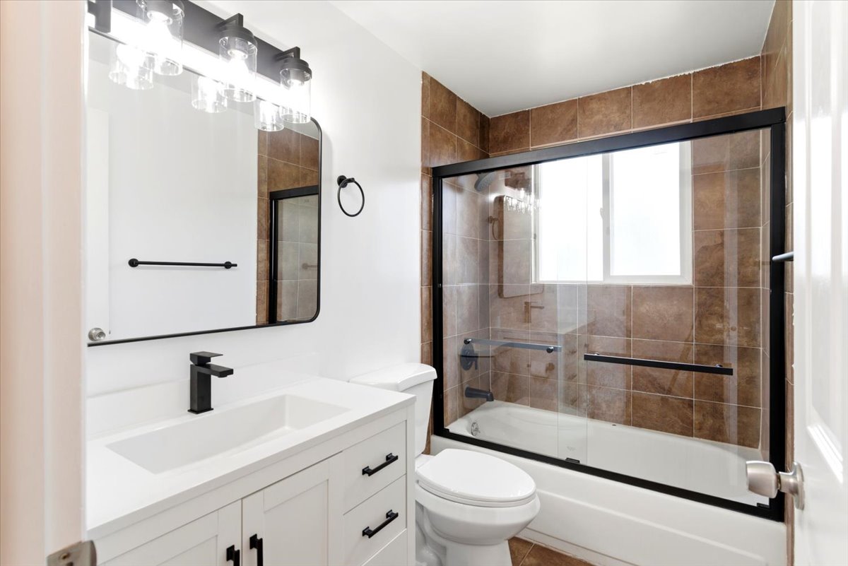 Bathroom featuring a white vanity with matte black hardware, a matte black faucet, a rectangular mirror, and a shower-tub combination with sliding glass doors and brown tile surround