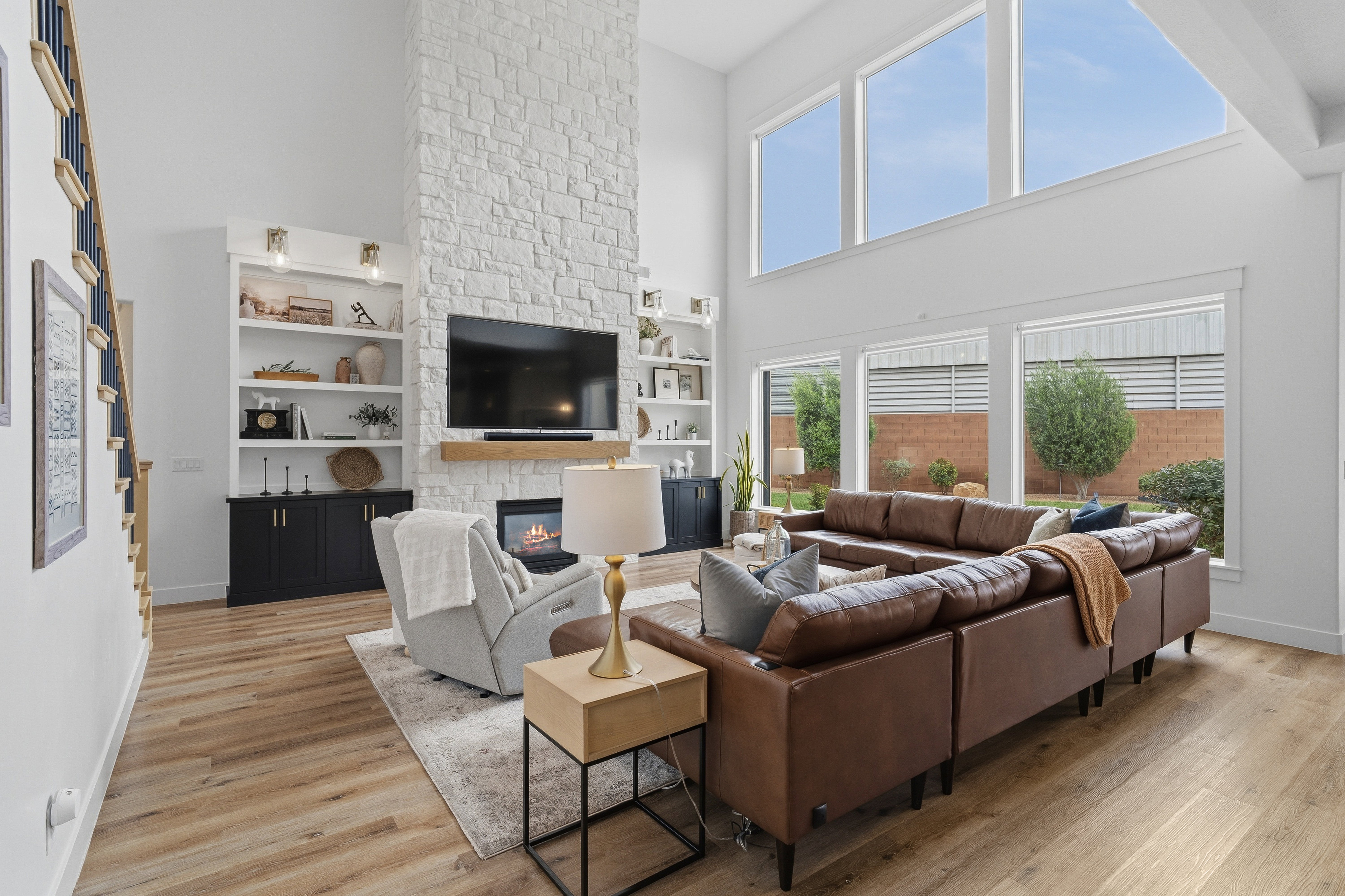 Vaulted living space featuring a stacked stone fireplace with wood mantel, large windows, wood-finish flooring, and built-in shelving with lower cabinetry.