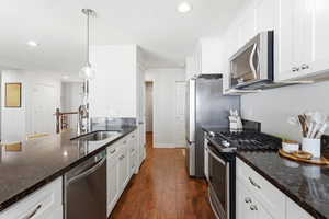 Modern kitchen featuring white cabinetry, dark stone countertops, stainless steel appliances, wood-finish flooring, and a central island with a sink and pendant lighting