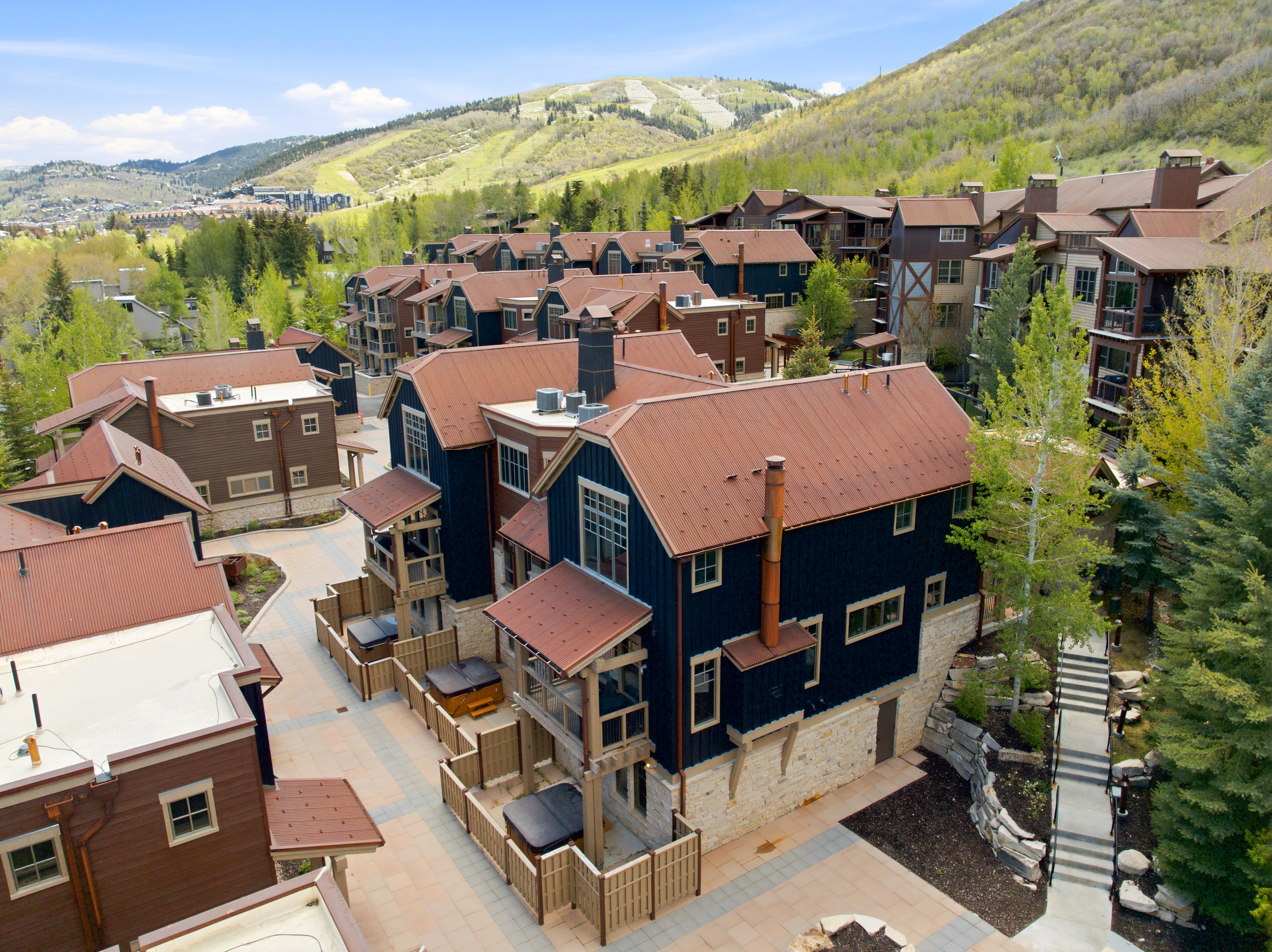 Community of multi-story townhomes featuring stone and dark siding exteriors, copper-toned metal roofs, individual patios with built-in spas, and a pedestrian pathway with stone retaining walls