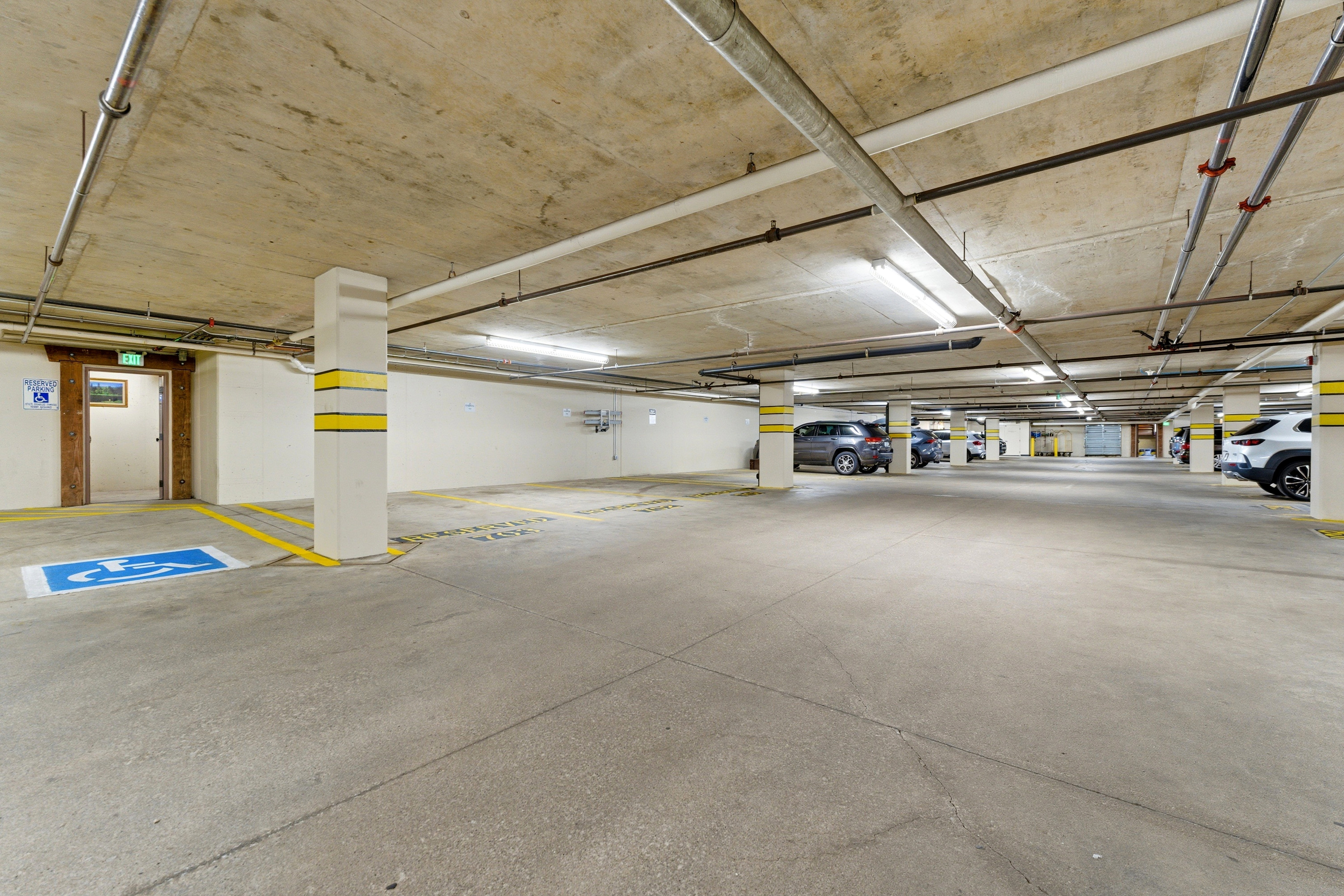 Spacious underground parking area featuring concrete flooring, exposed pipework, and fluorescent lighting