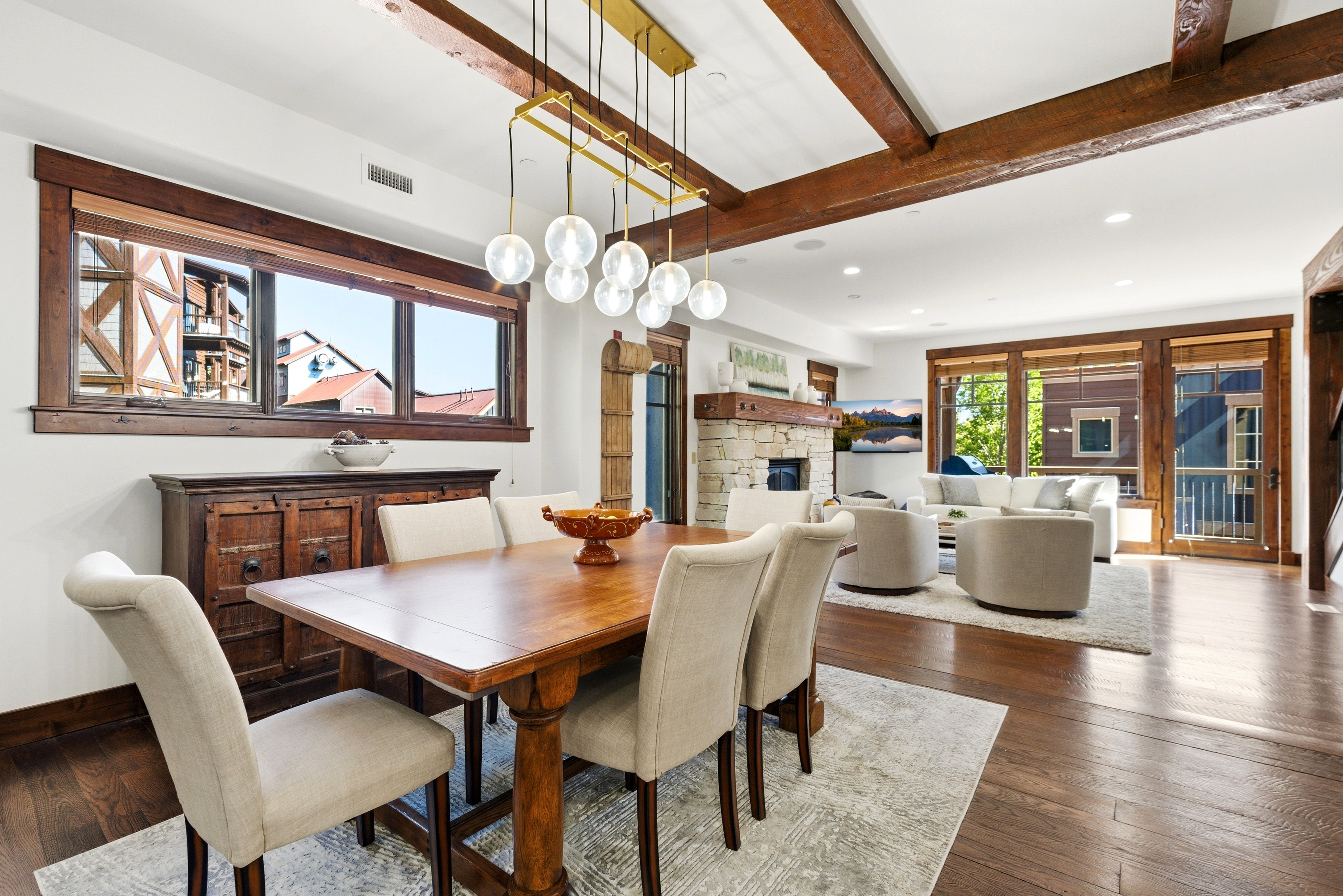 Open-concept living space featuring wood-finish flooring, exposed ceiling beams, a stone-clad fireplace, and large windows with wood trim