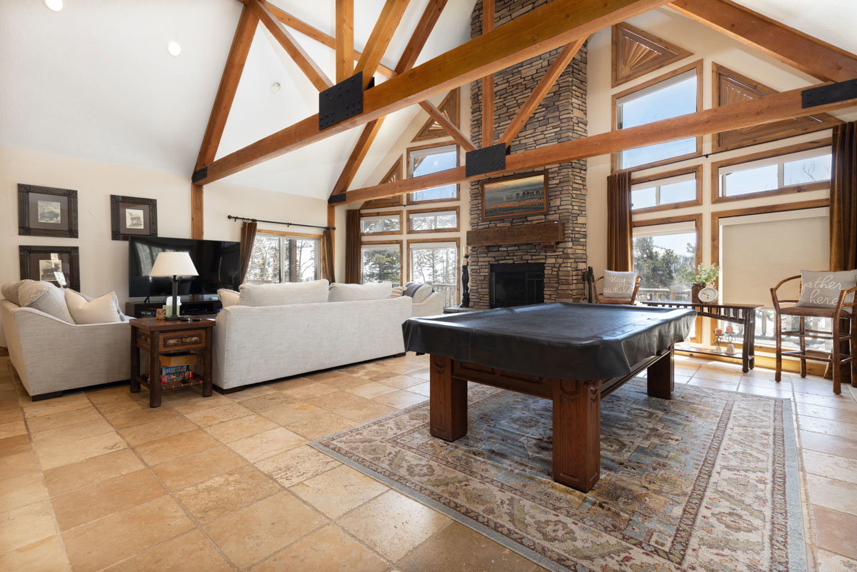 Vaulted ceilings with exposed wood beams, stone-faced fireplace with wood mantel, large windows with natural wood trim, and tile flooring