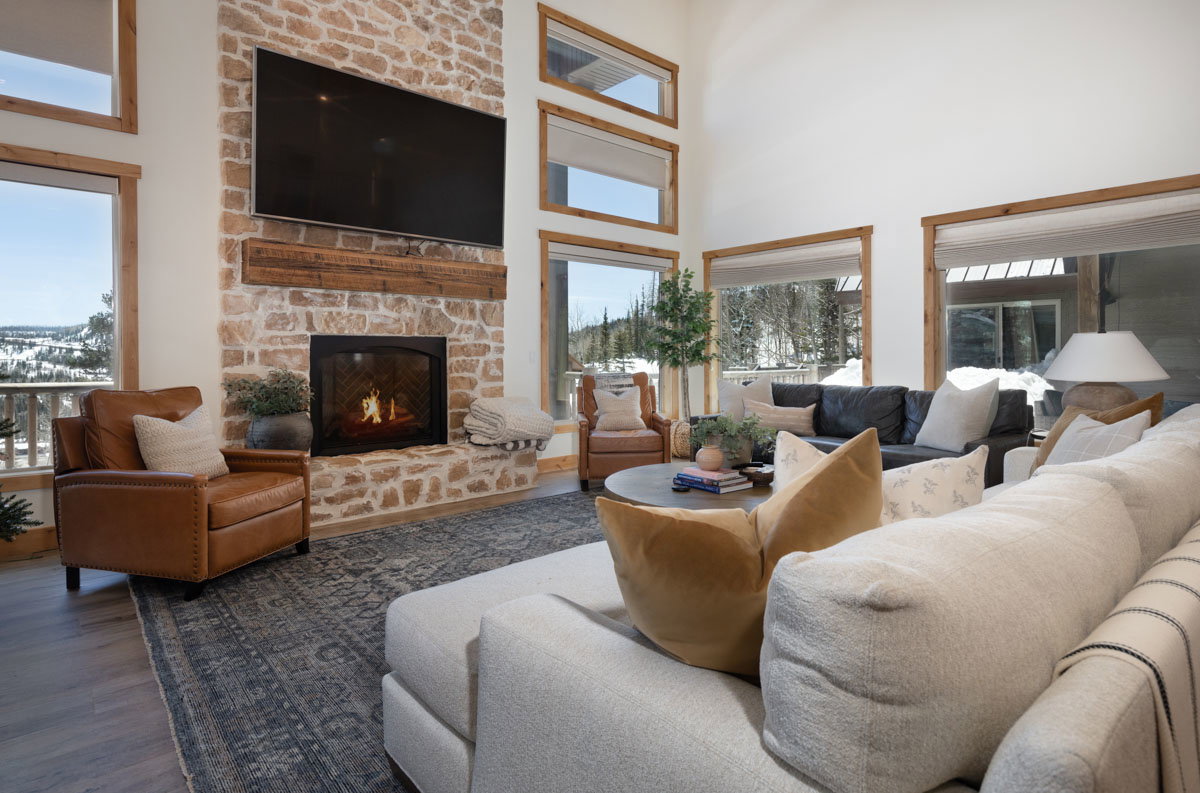 Living area featuring a stone-clad fireplace with a wood mantel, high ceilings, large windows with wood trim, and wood-finish flooring