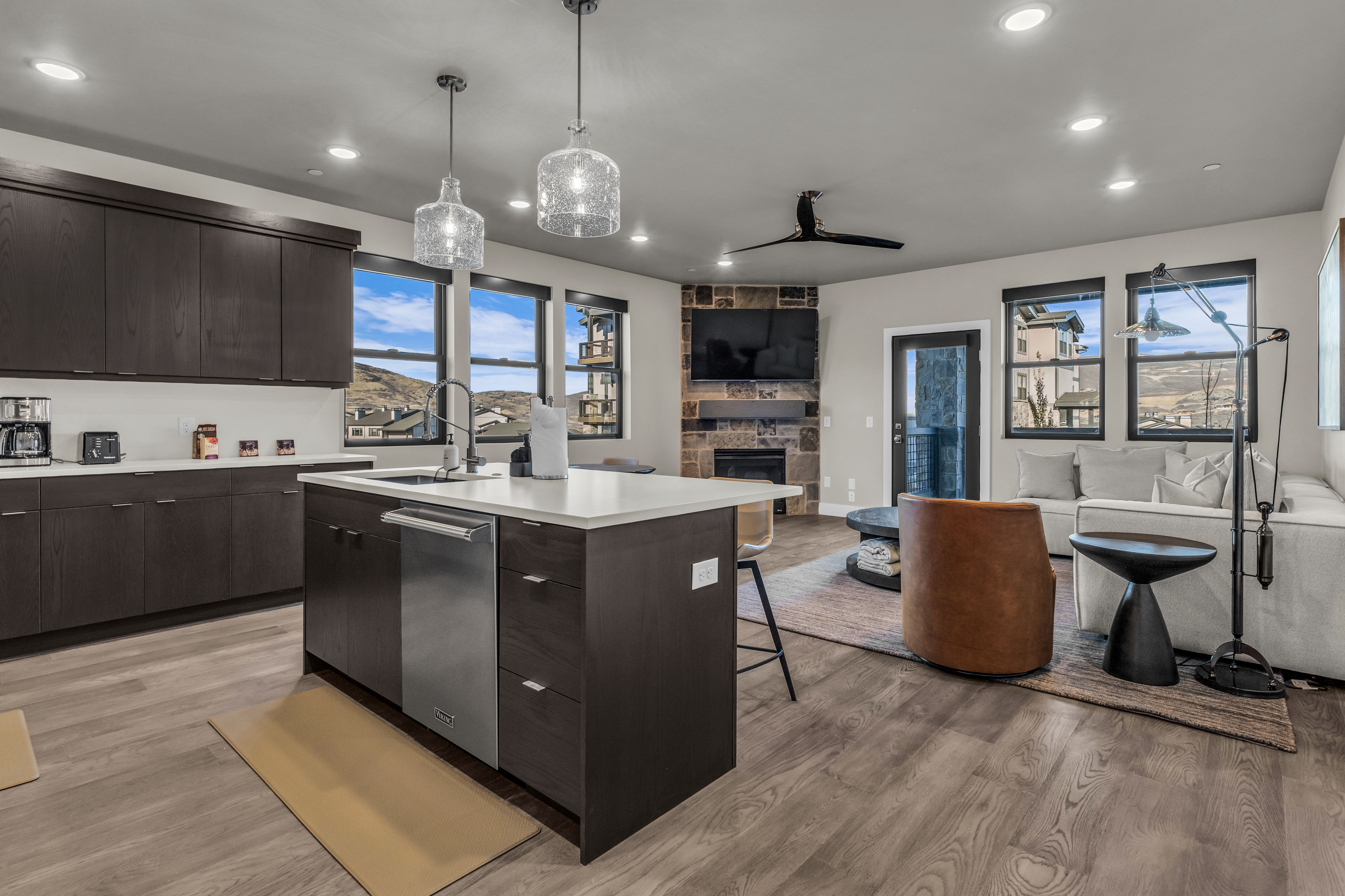 Modern kitchen and living area featuring dark wood-finish cabinetry, a central island with a white countertop and integrated stainless steel dishwasher, and wood-finish flooring