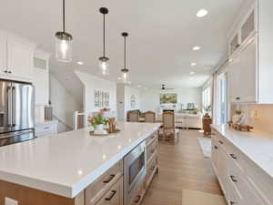 Spacious kitchen featuring a large island with built-in microwave, light-toned wood-finish flooring, white shaker cabinetry, stainless steel appliances, and sleek pendant lighting
