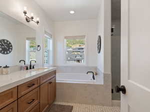Bathroom featuring a dual vanity with a light-toned countertop and wood-finish cabinetry, a drop-in soaking tub with a tiled surround, a walk-in shower with a glass enclosure, and hexagonal mosaic tile flooring