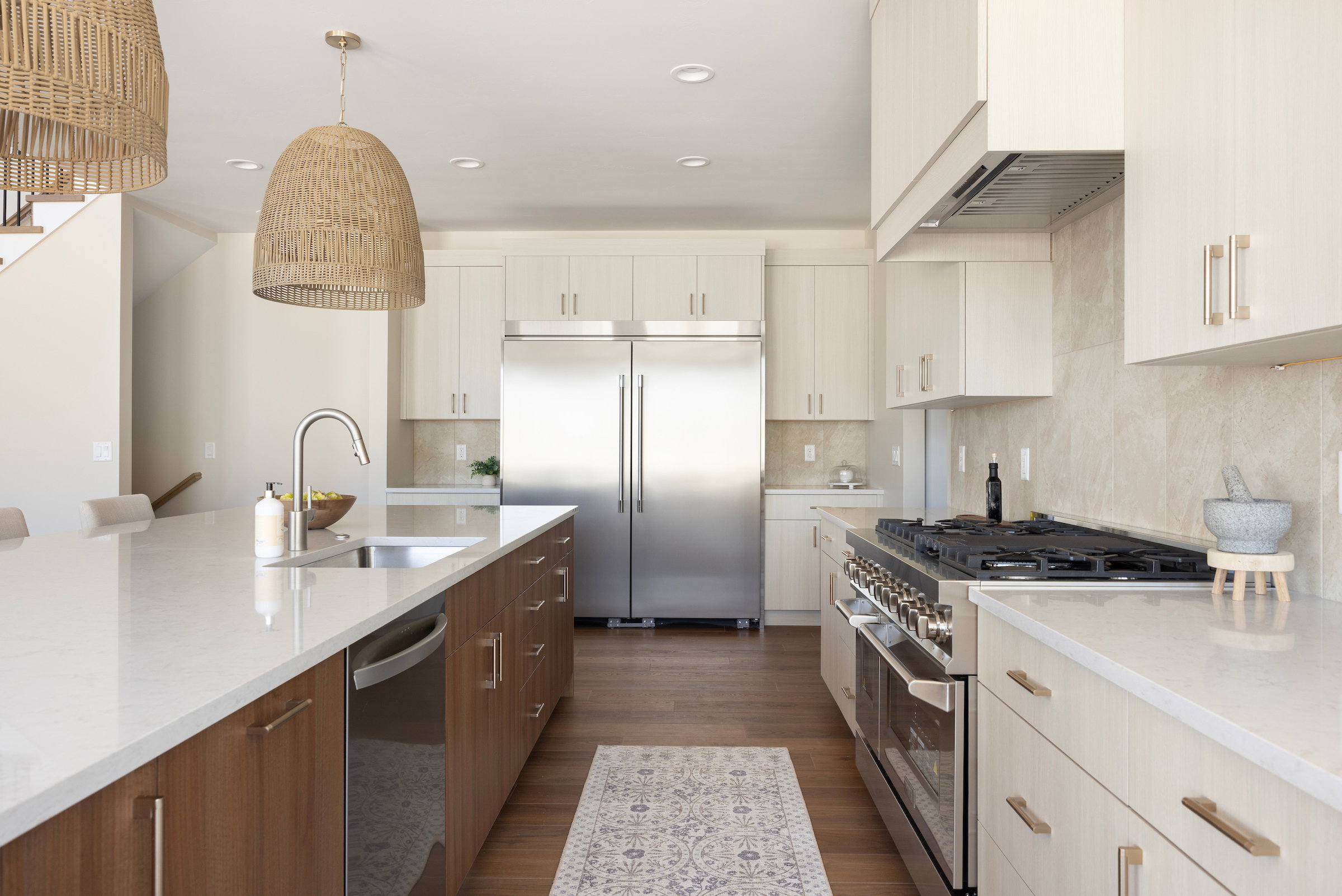 Gourmet kitchen featuring a large central island with an undermount sink, stainless steel appliances, light-toned upper cabinetry, wood-finish base cabinetry, and a full-height backsplash