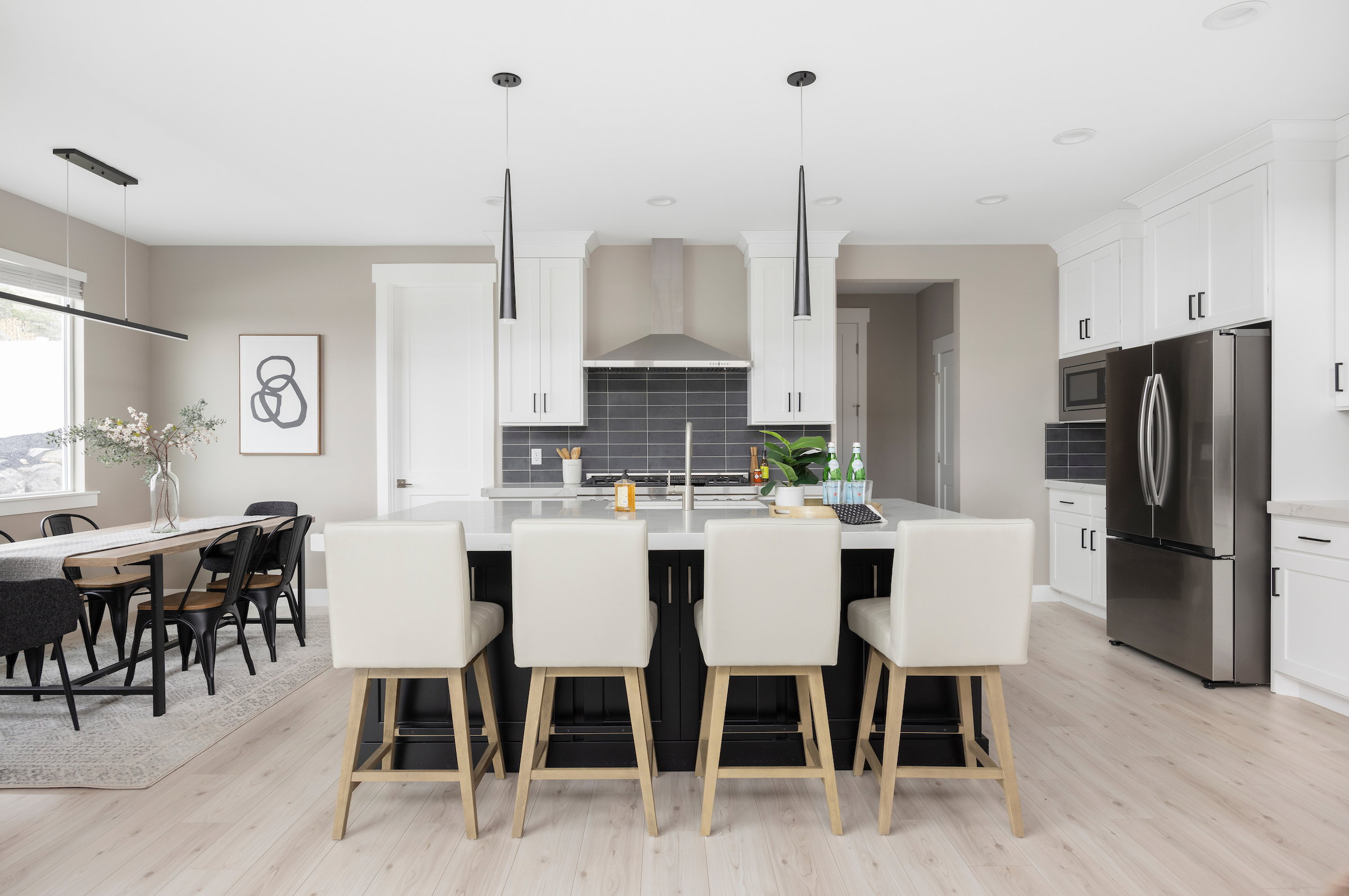 Open-concept kitchen featuring a large central island with seating, stainless steel appliances, white cabinetry with dark hardware, a dark subway tile backsplash, and light wood-finish flooring