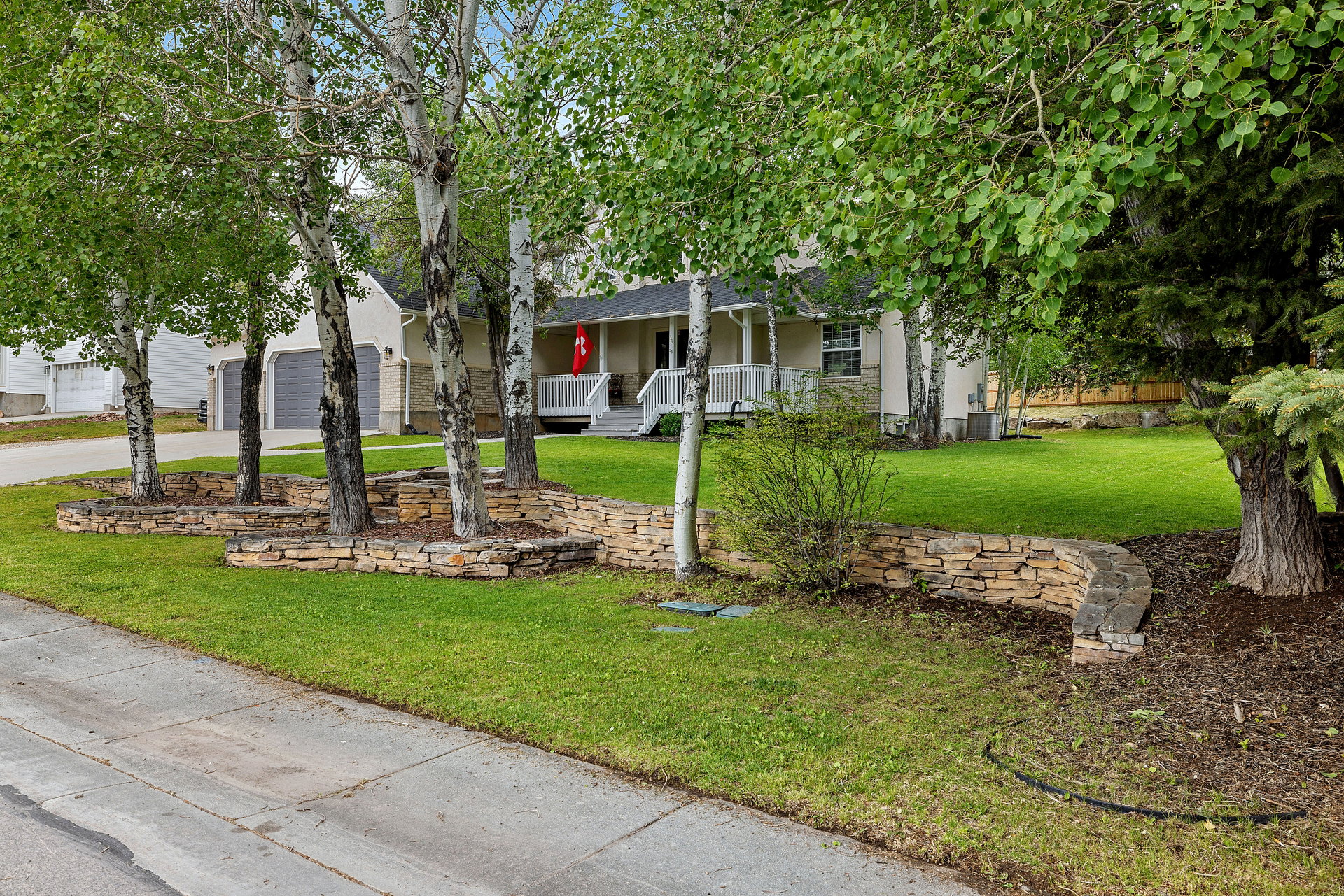 Front yard landscaping featuring multiple mature trees, stone retaining walls, and a manicured lawn