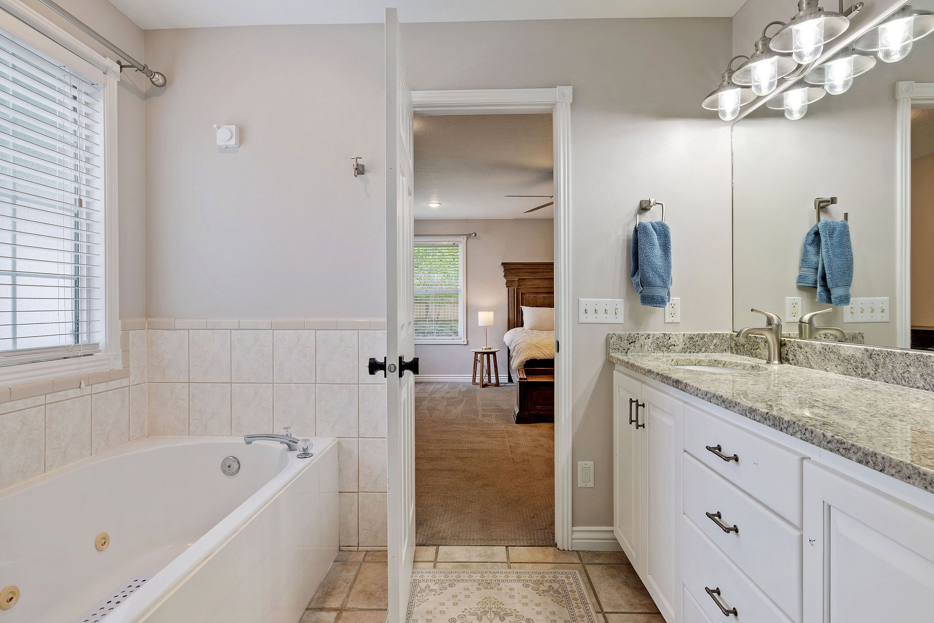 Bathroom featuring a built-in jetted tub, a window with horizontal blinds, tiled flooring, a double vanity with a light-colored granite countertop, and a large mirror with overhead lighting