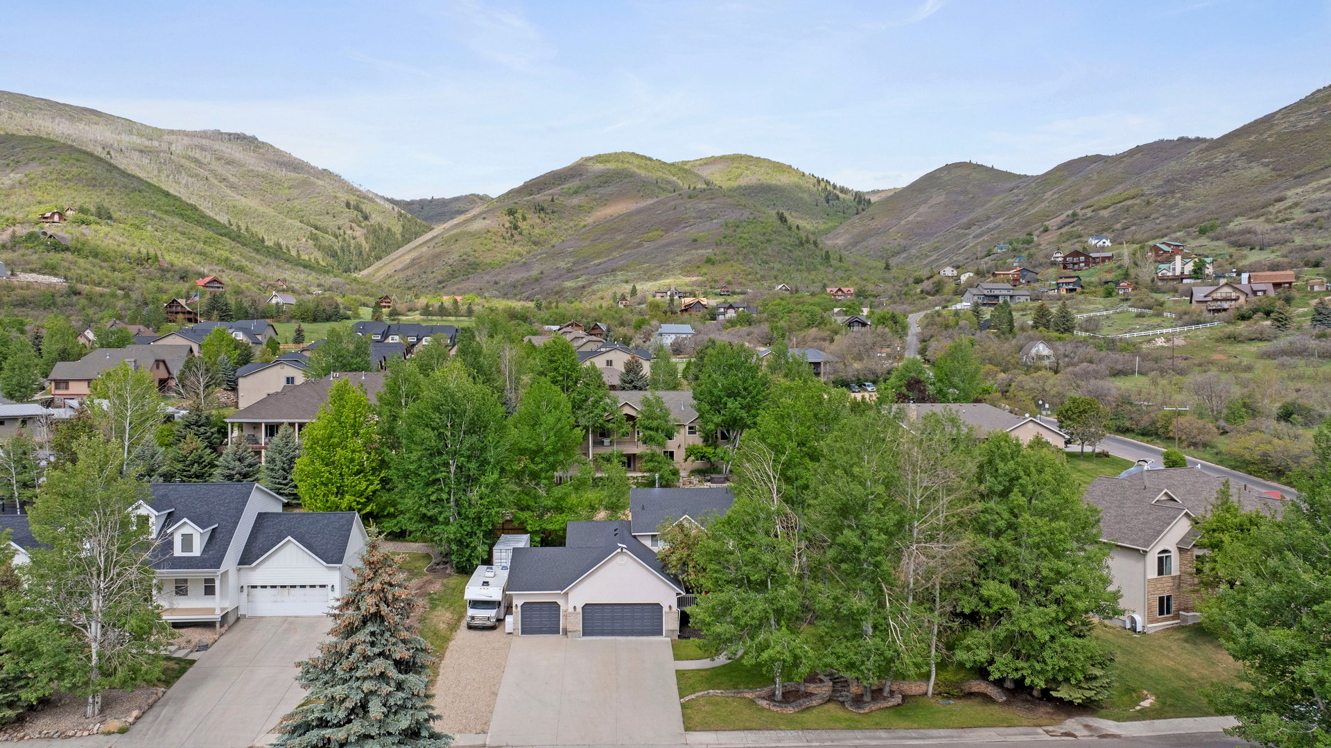 Mountainous terrain surrounds a residential area featuring multiple homes with varied architectural styles, mature trees, and paved driveways