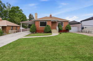 Brick exterior residence featuring a front lawn, paved driveway, covered carport, and mature shrubs
