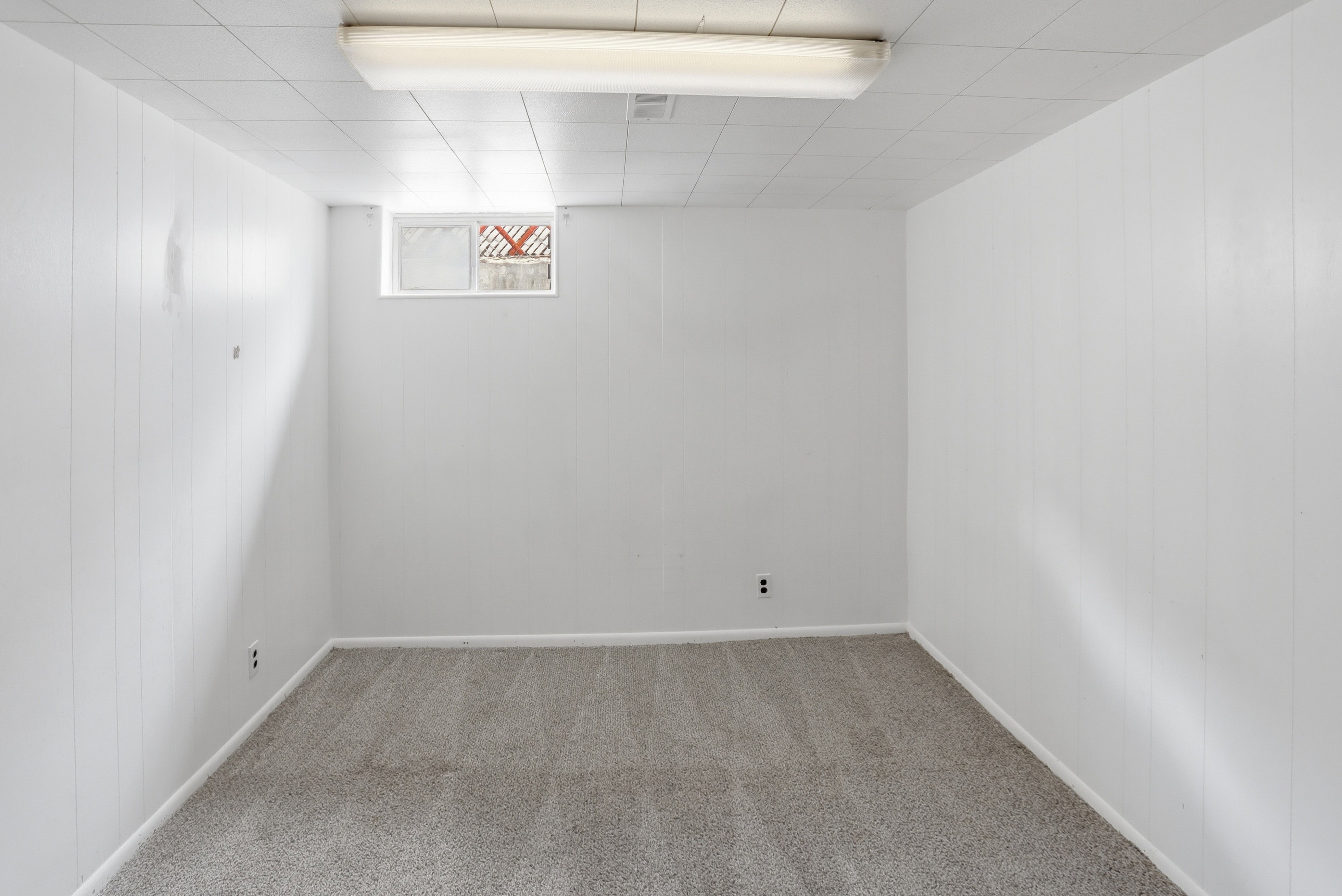 Spacious room featuring light gray carpeting, crisp white paneled walls, a drop ceiling with integrated lighting, and a single window