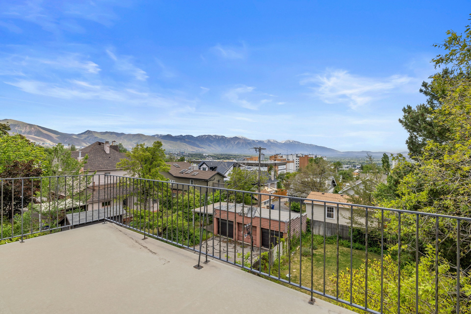 Rooftop patio featuring a concrete surface and black metal railing, overlooking urban surroundings with distant mountains