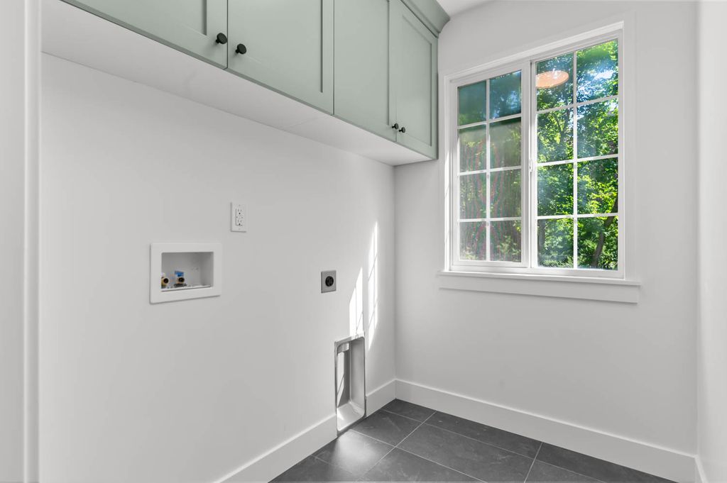 Laundry room featuring light green upper cabinetry with dark hardware, a large window with gridded panes, dark tile flooring, and white baseboards