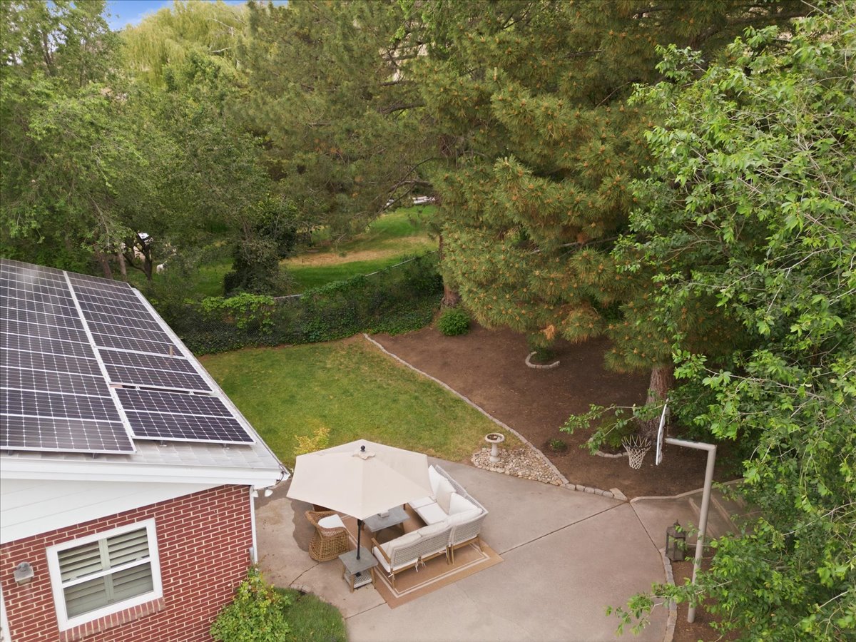 Brick exterior with solar panels, a paved patio, and a basketball hoop, surrounded by mature trees and a manicured lawn